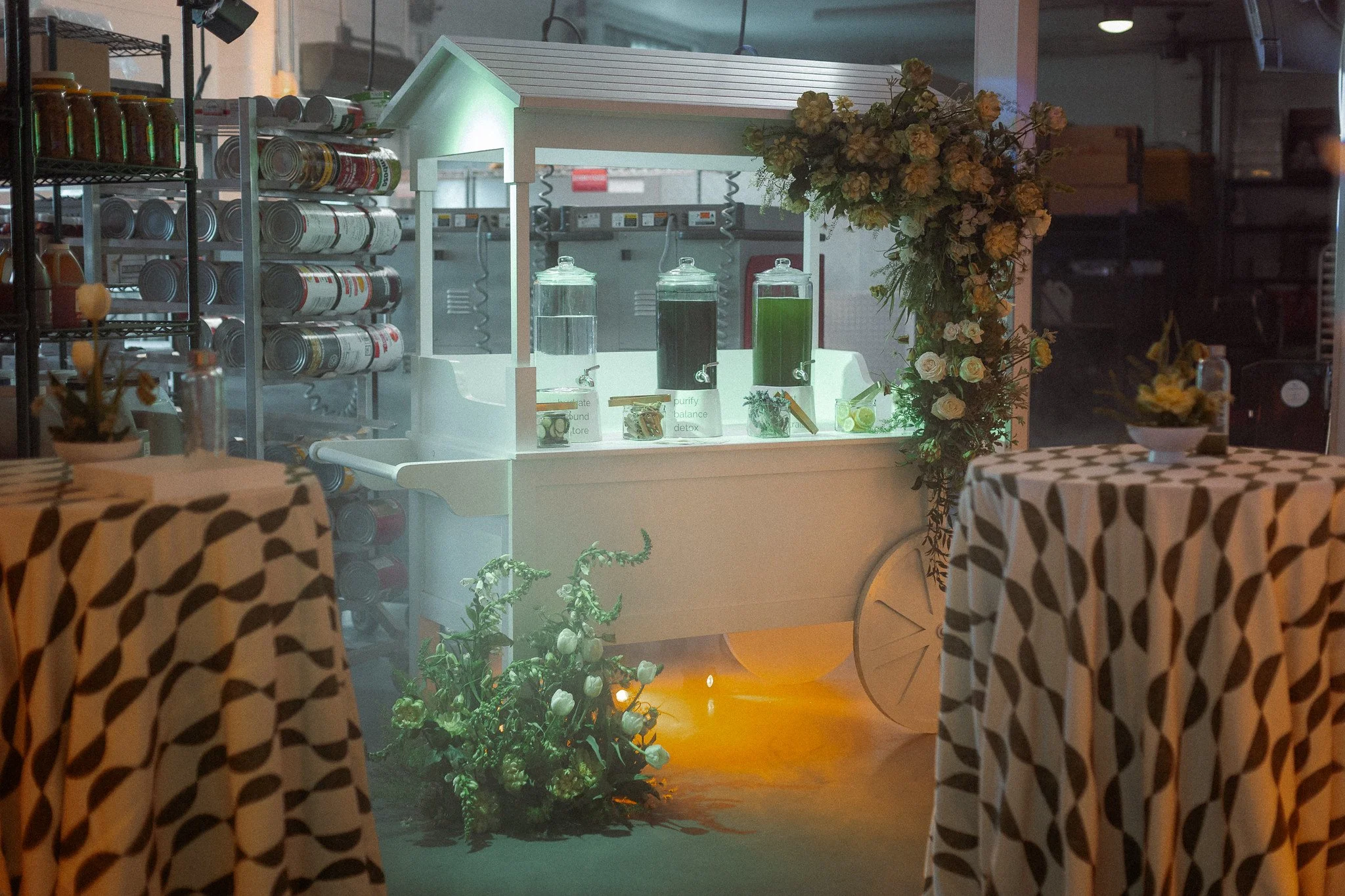 A food service or beverage stand decorated with flowers, with tables covered in black and white patterned tablecloths in the foreground. The stand features three glass containers with green drinks, a small sign, and a flower arrangement on the right.