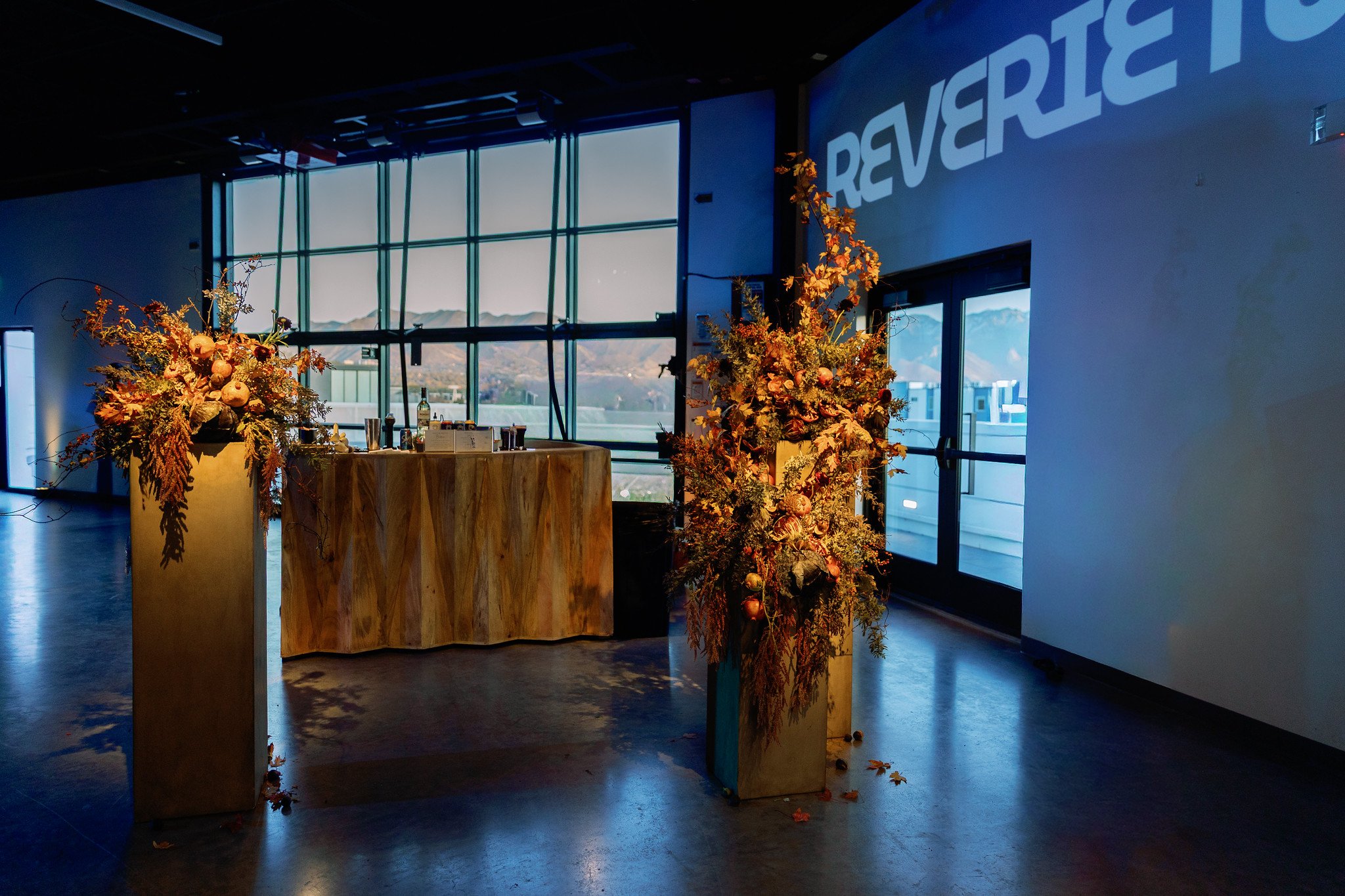 Interior of a modern building with large windows, autumnal floral arrangements on wooden stands, and a partial word 'REVERS' projected on the wall