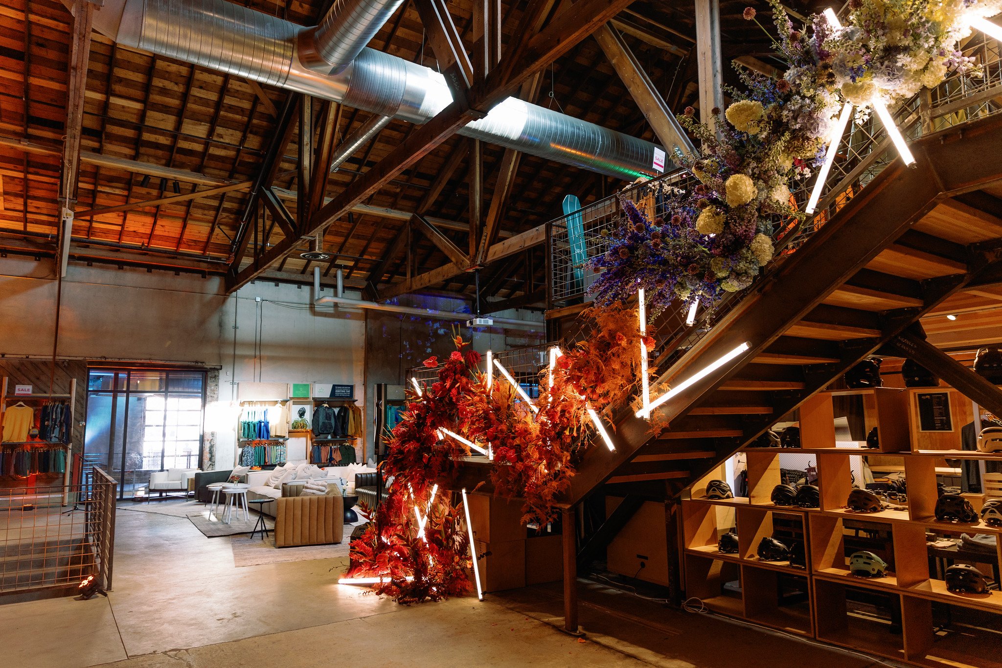Interior of a modern building with exposed wooden beams, ventilation ducts, a staircase decorated with flowers, sneakers, helmets, and various seating and retail areas.