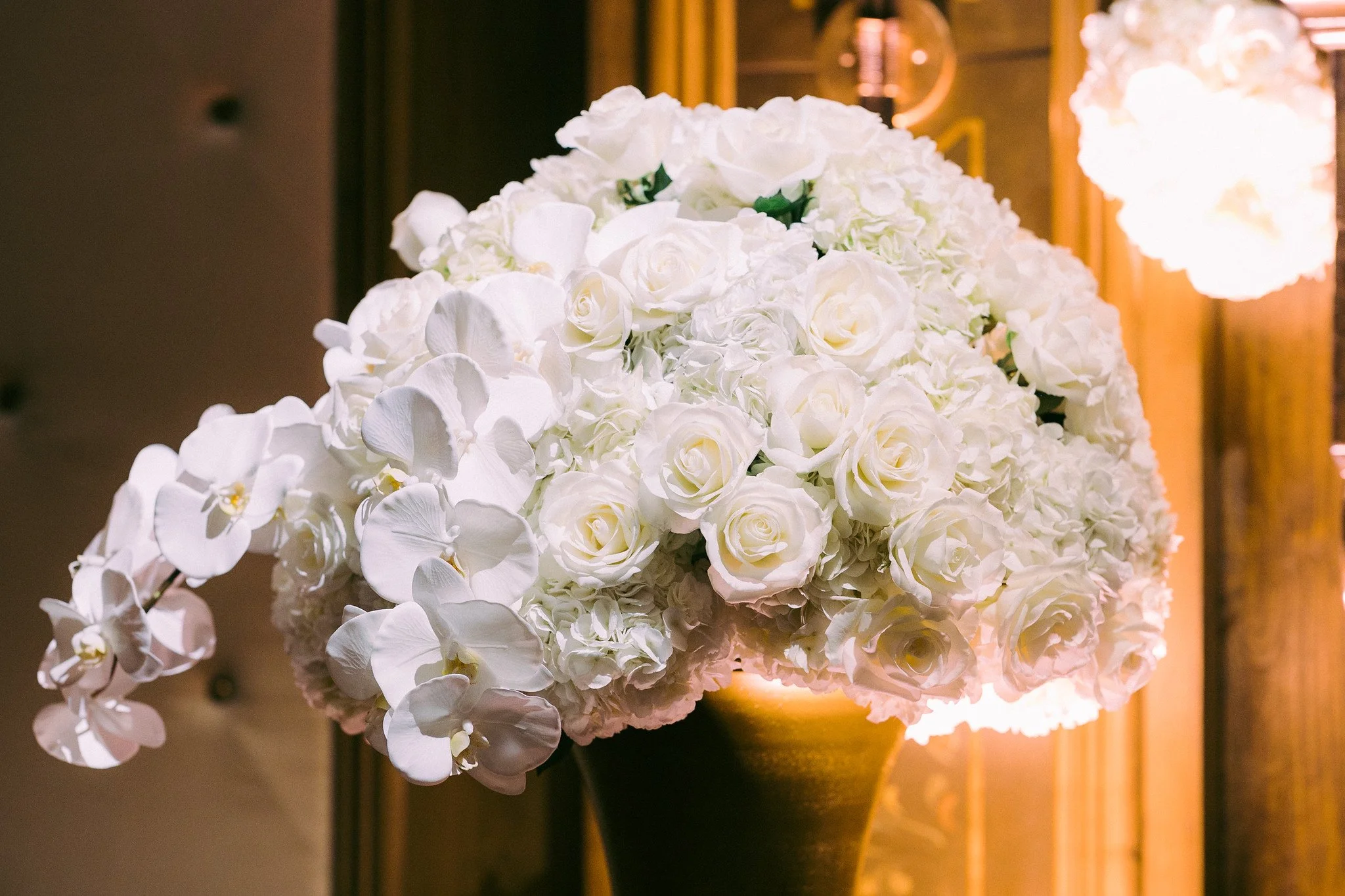 A bouquet of white roses, orchids, and hydrangeas placed on a wooden table, with a warm lamp illuminating the background.