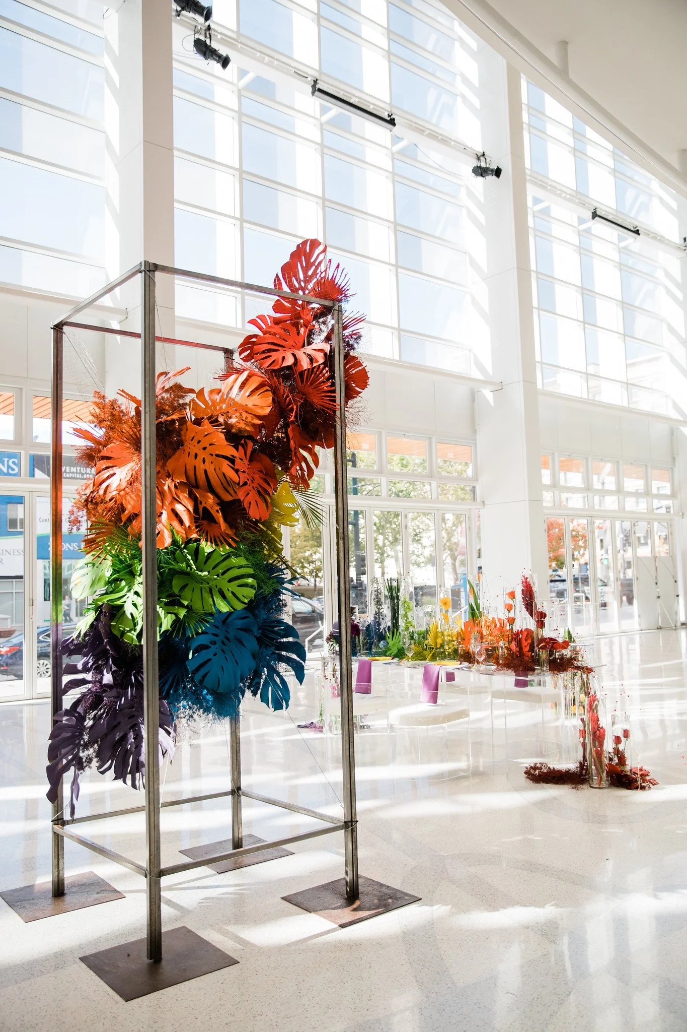 A colorful display of tropical leaves in shades of orange, purple, blue, and green arranged on a metal frame inside a bright, modern building with large windows.