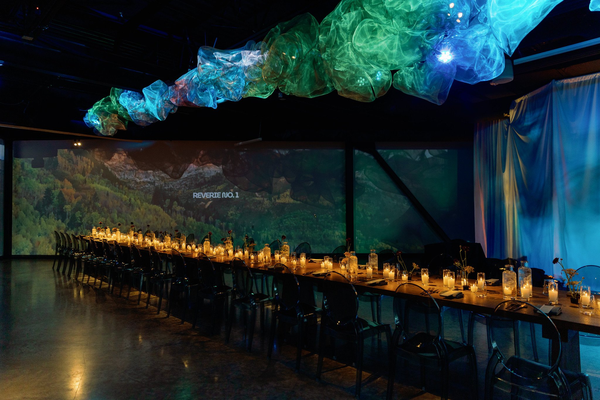 A decorated banquet hall with a long table set with candles and elegant glass bottles, large colorful fabric decorations hanging from the ceiling, and a large screen displaying a scenic landscape with the words "Reverte No.1." in the background.