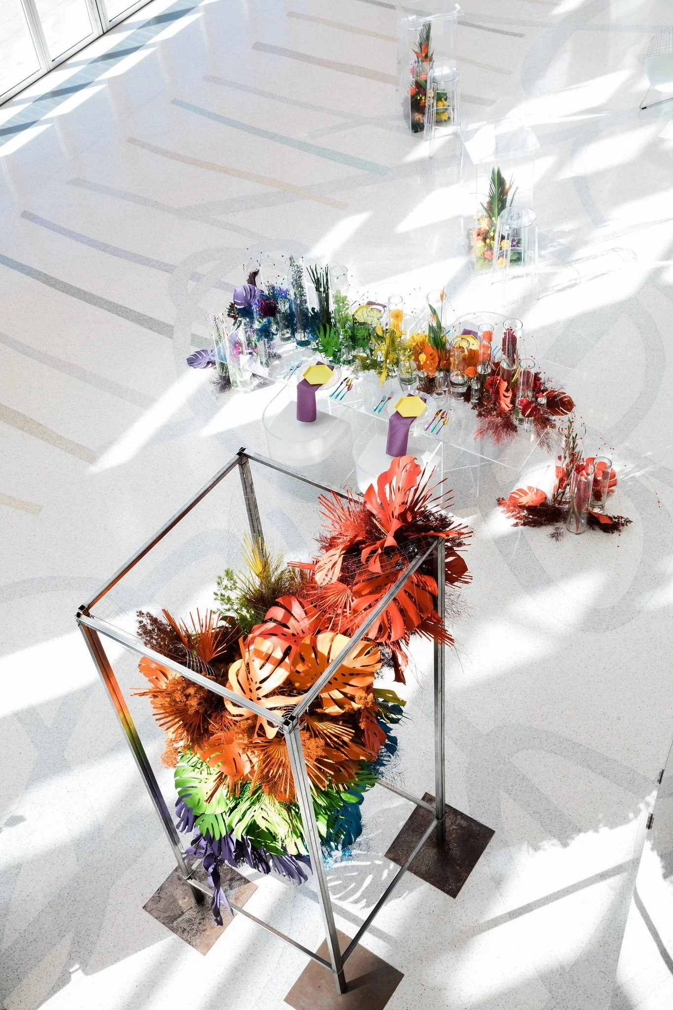 Colorful floral arrangements with tropical and exotic plants displayed on transparent acrylic stands inside a bright, modern space with white flooring and large windows casting shadows.