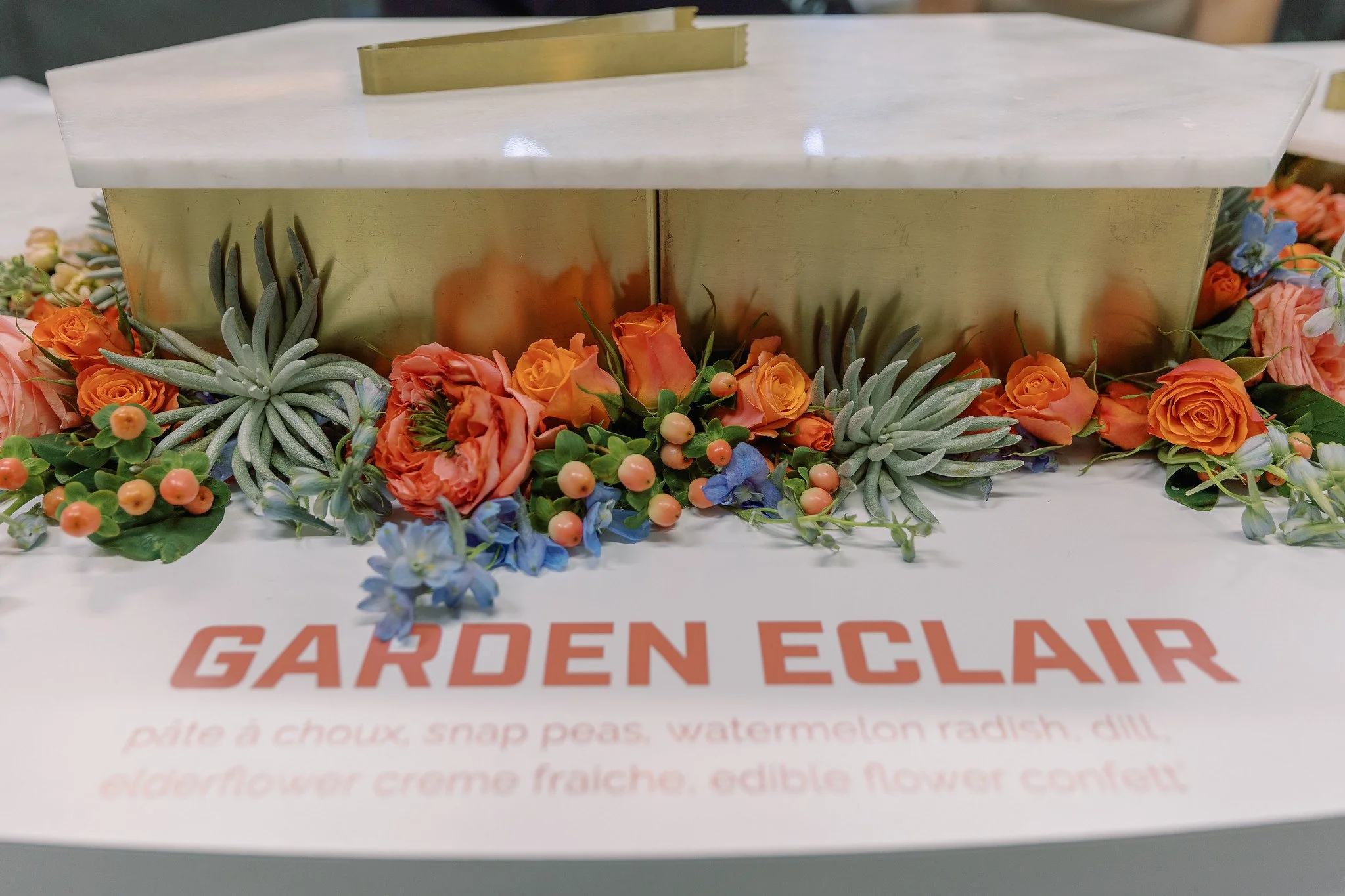 A floral arrangement with orange roses, blue and purple flowers, and green foliage surrounds a gold-colored rectangular box with a white marble lid, placed on a white surface with partially visible orange and red text.