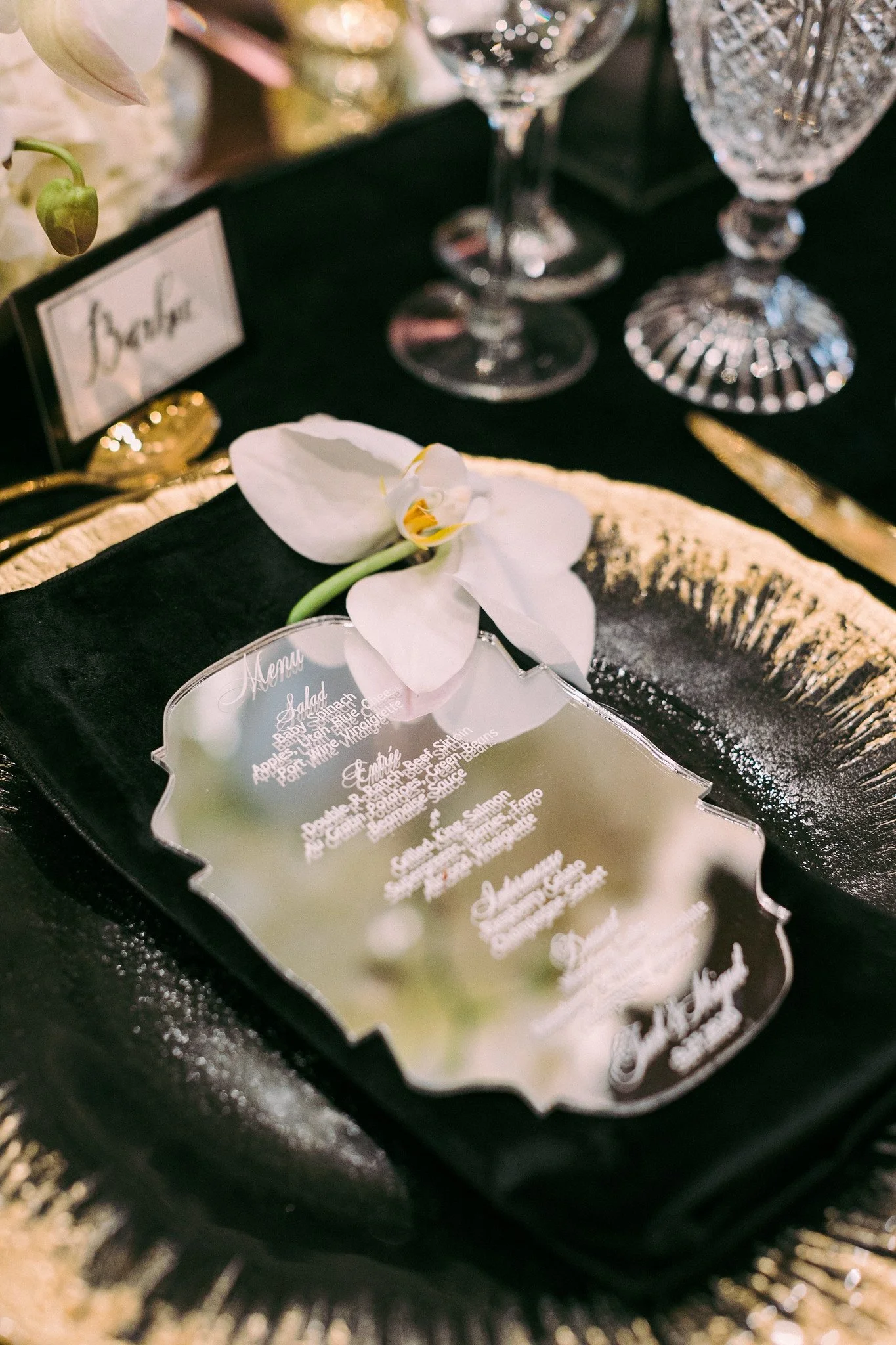 Close-up of a dessert or food item on a menu with a white orchid flower on top, on a black tray with a reflective surface, and glasses in the background.