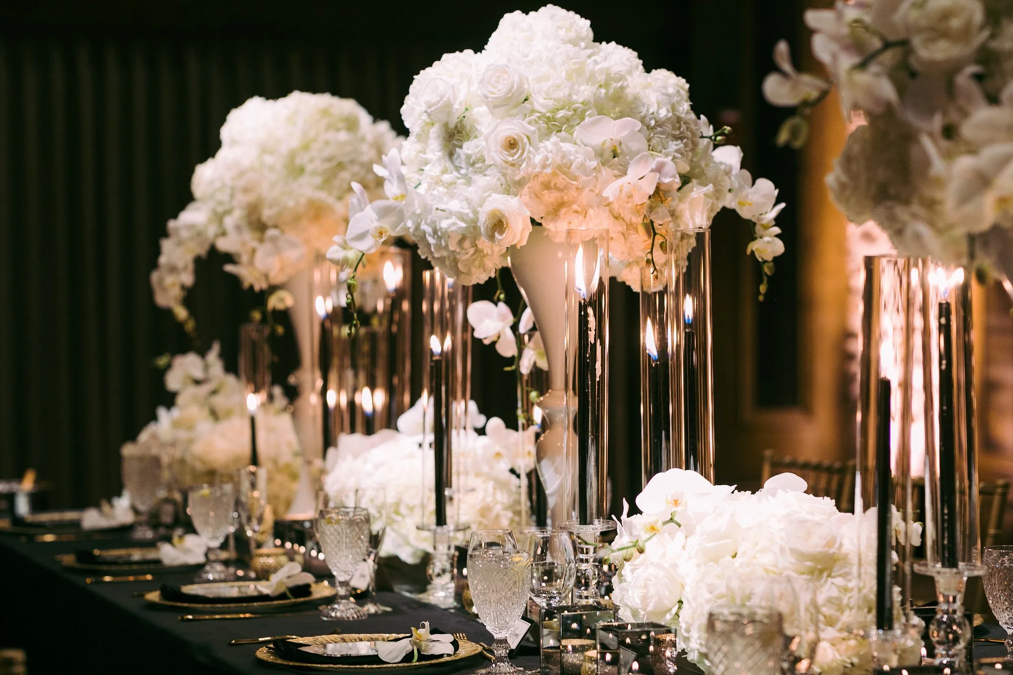 Elegant event table decorated with tall white floral arrangements, surrounded by candles and glassware.