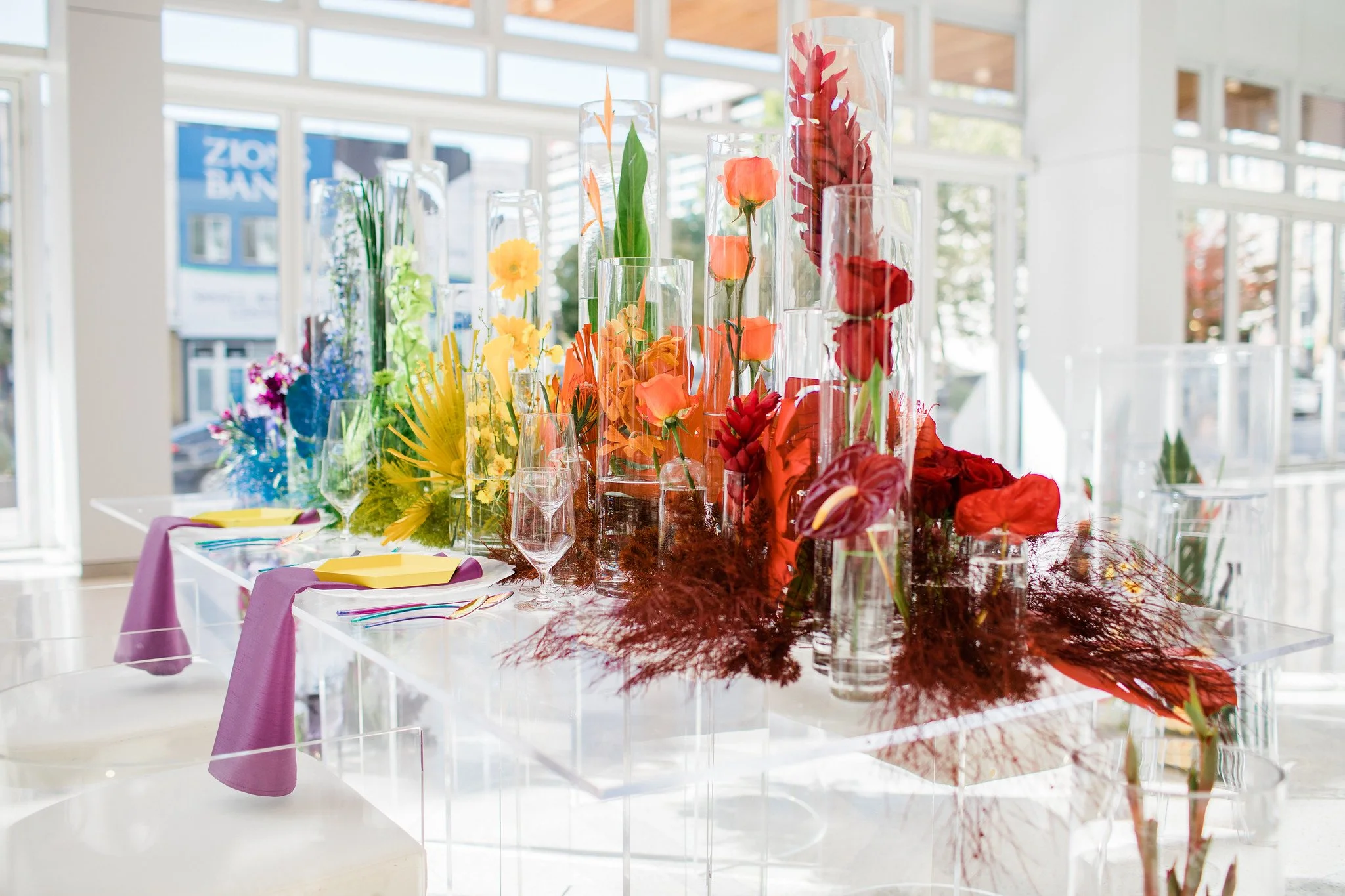 Table decorated with a colorful floral centerpiece in tall glass vases, with purple and yellow napkins on white tablecloth, inside a bright room with large windows.