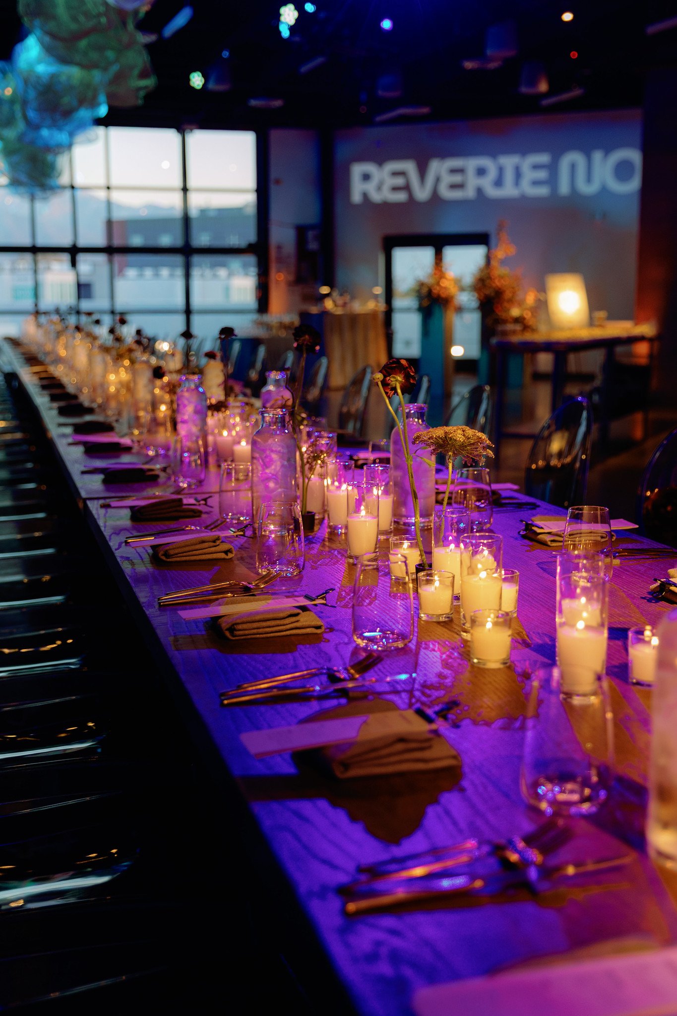 Long dinner table decorated with lit candles, vases with flowers, and glassware in a dimly lit room with large windows and purple lighting.