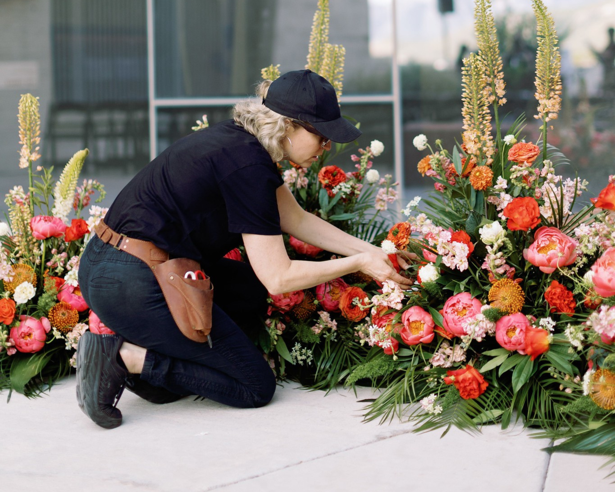 A woman wearing a black cap, black shirt, and black boots is kneeling and arranging a large colorful flower display with pink, orange, and white flowers, in an outdoor setting in front of a glass building.
