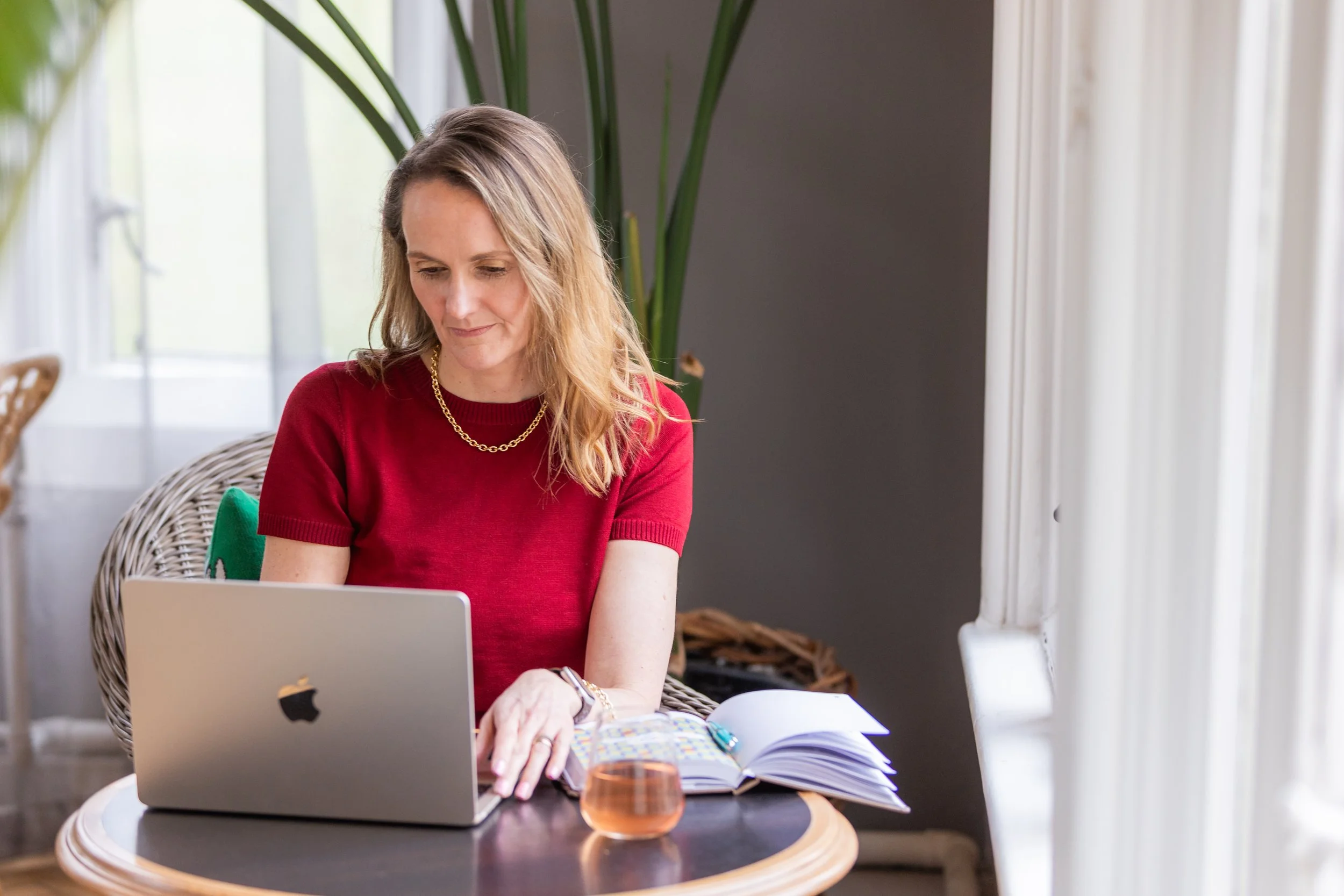 A woman with blonde hair wearing a red shirt and gold necklace working at a desk with a laptop, notebooks, and a glass of pink beverage in a bright room with large windows and green plants.