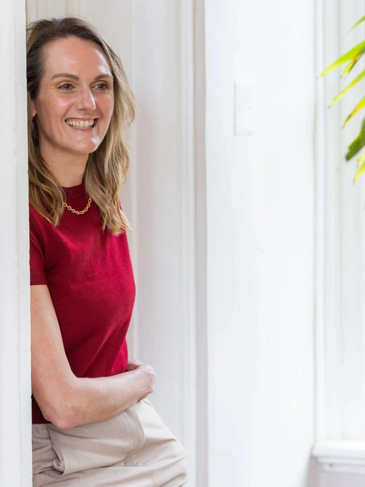 A woman with shoulder-length blonde hair, wearing a red shirt and beige pants, smiling while leaning against a white wall in a brightly lit room with a plant in the background.