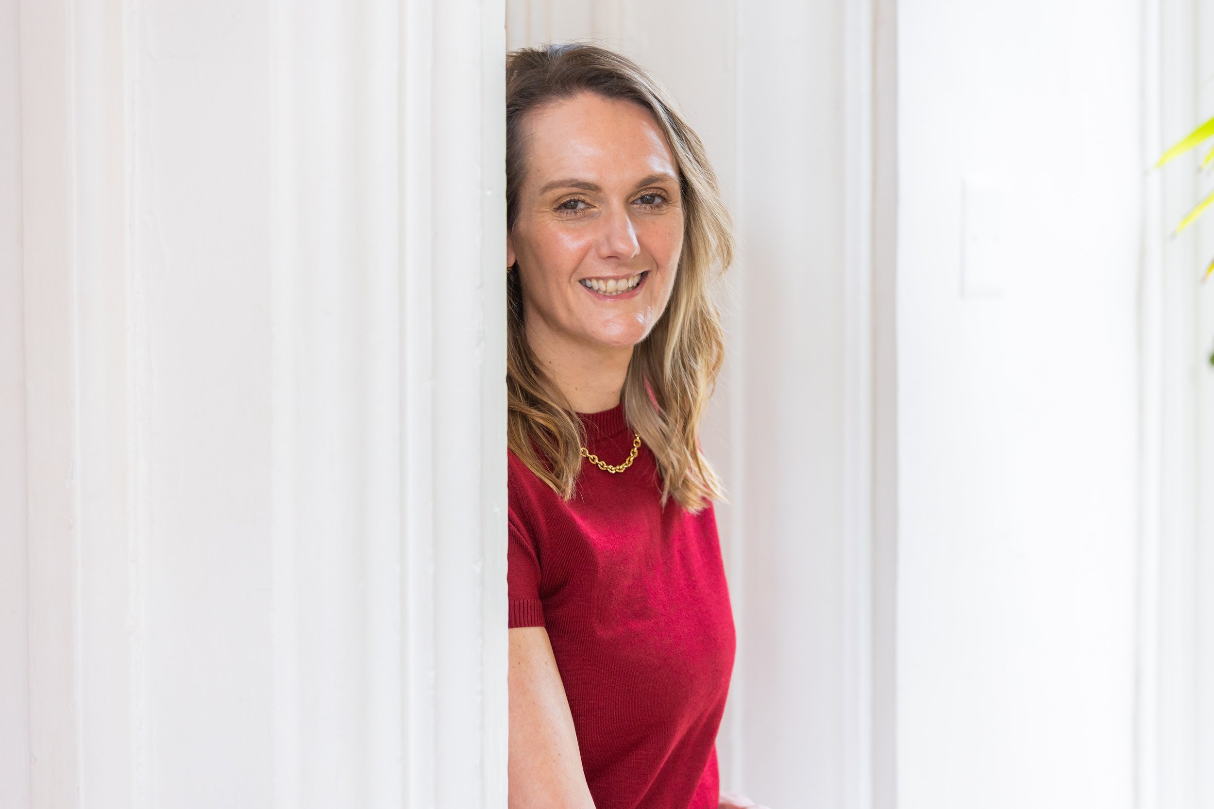A woman with light brown hair wearing a red dress and gold necklace, peeking out from behind a white curtain, smiling at the camera.