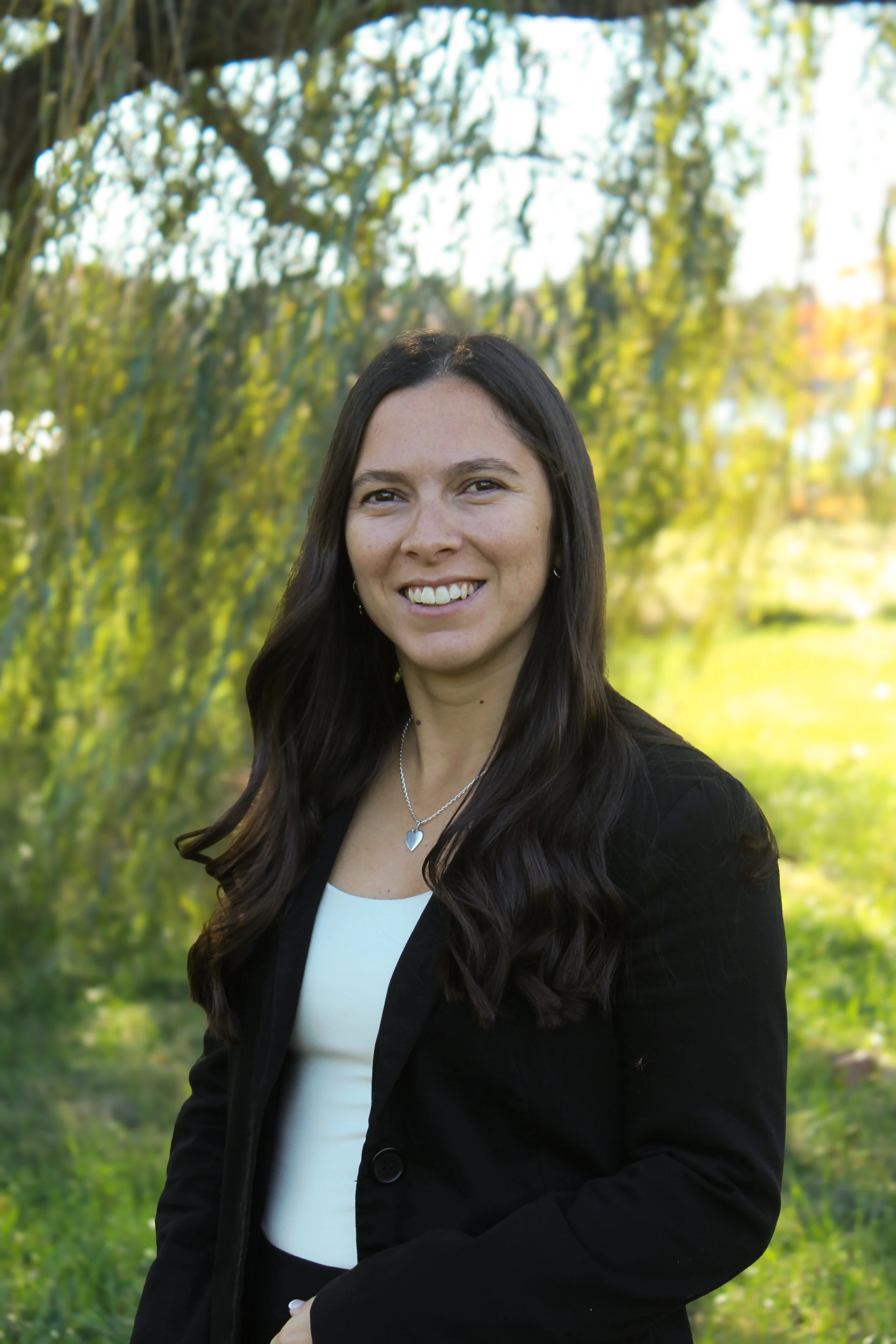 A woman with long dark hair, wearing a black blazer and a white top, smiling outdoors with greenery and sunlight in the background.