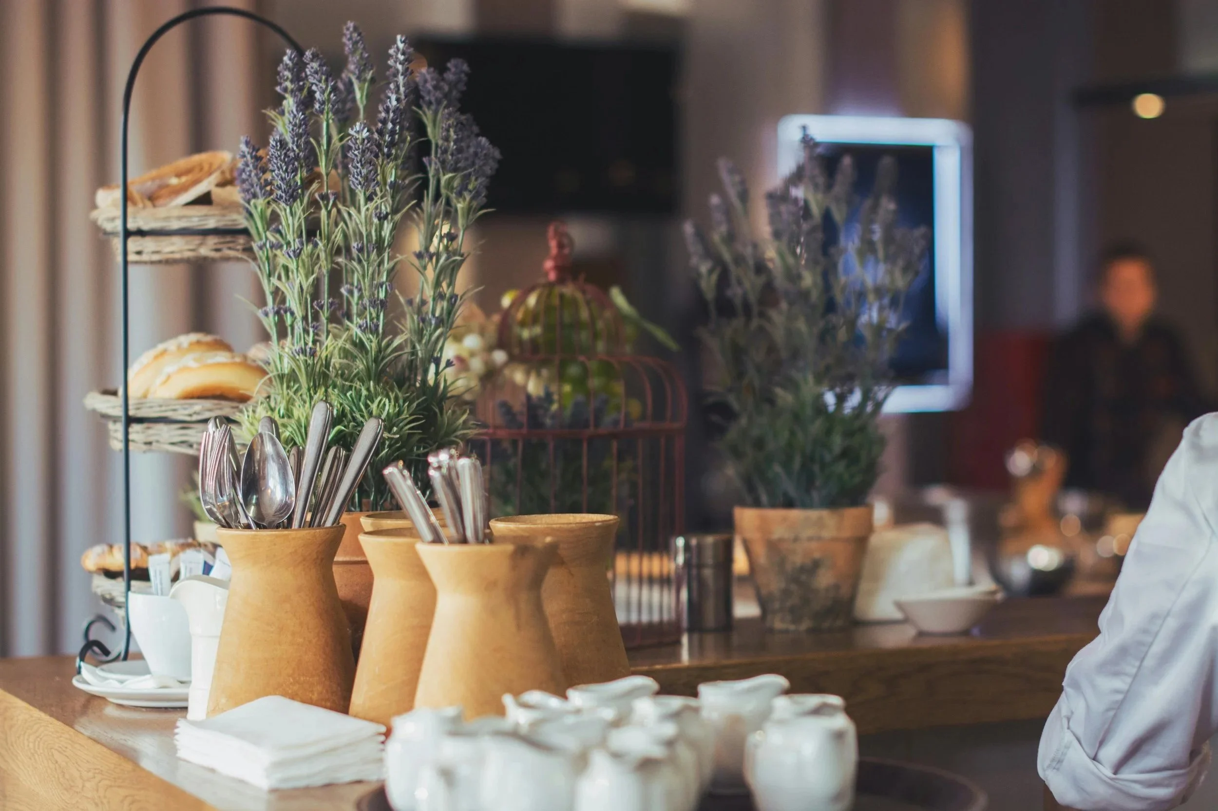 Arrangement of purple flowers, possibly lavender, in small clay pots on a wooden counter, with various bowls and utensils nearby, in a cozy indoor setting.