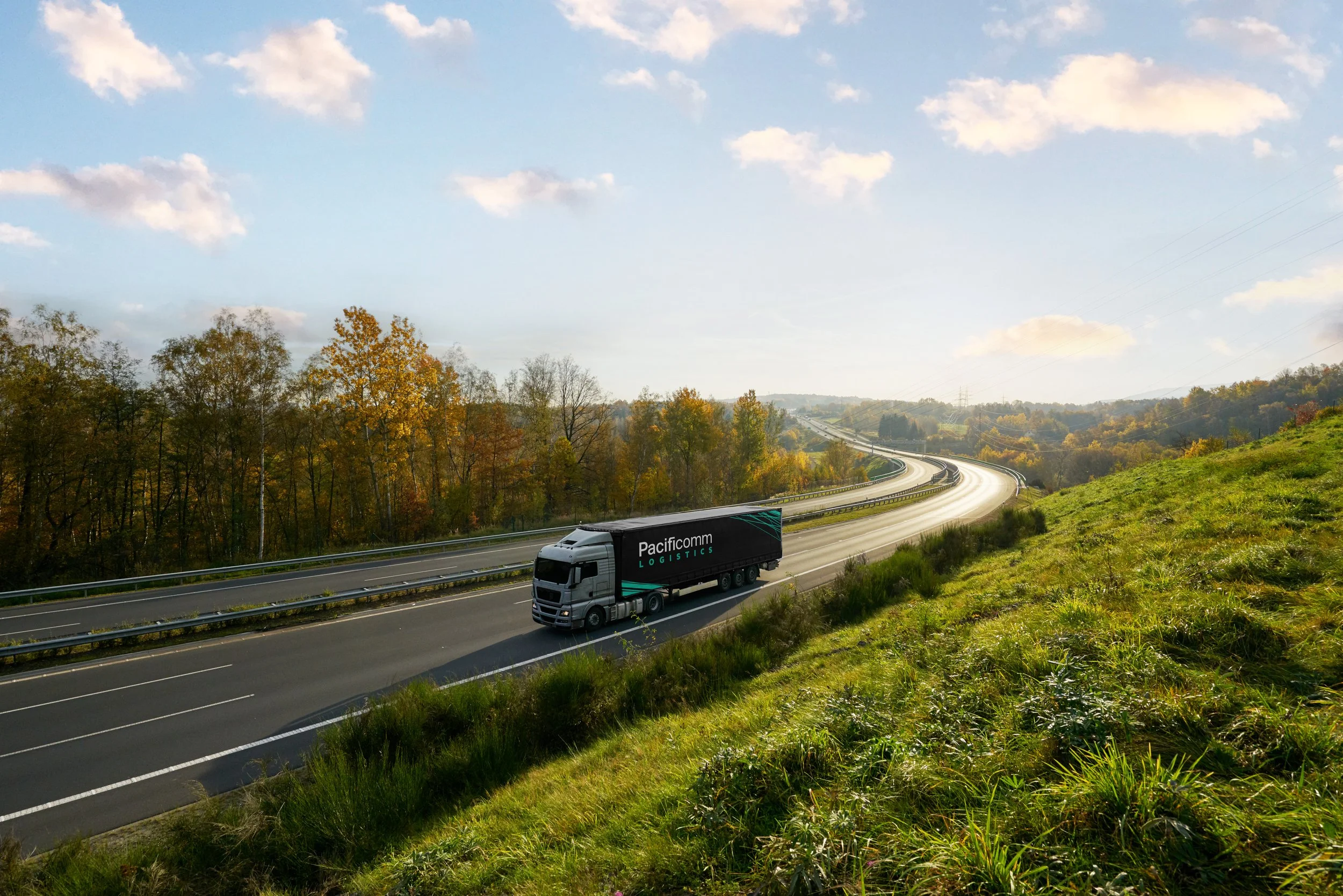 A highway with a truck labeled 'Pacificomm Logistics' driving on the right lane. The scene features a grassy slope on the right and a wooded area with autumn trees on the left. The sky is blue with scattered clouds.