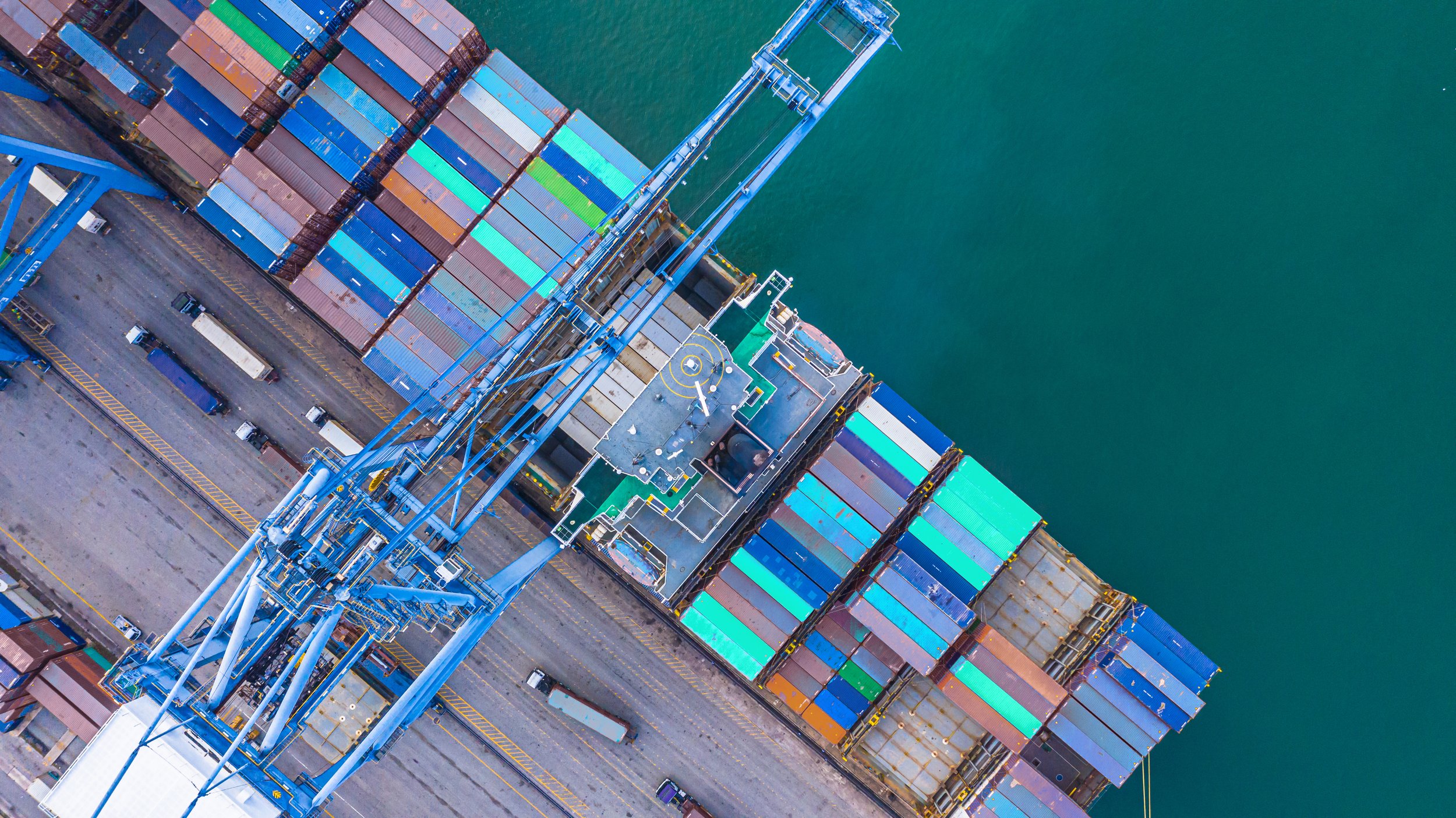 Aerial view of a large cargo ship at a port, with colorful shipping containers stacked on deck and a blue crane overhead.