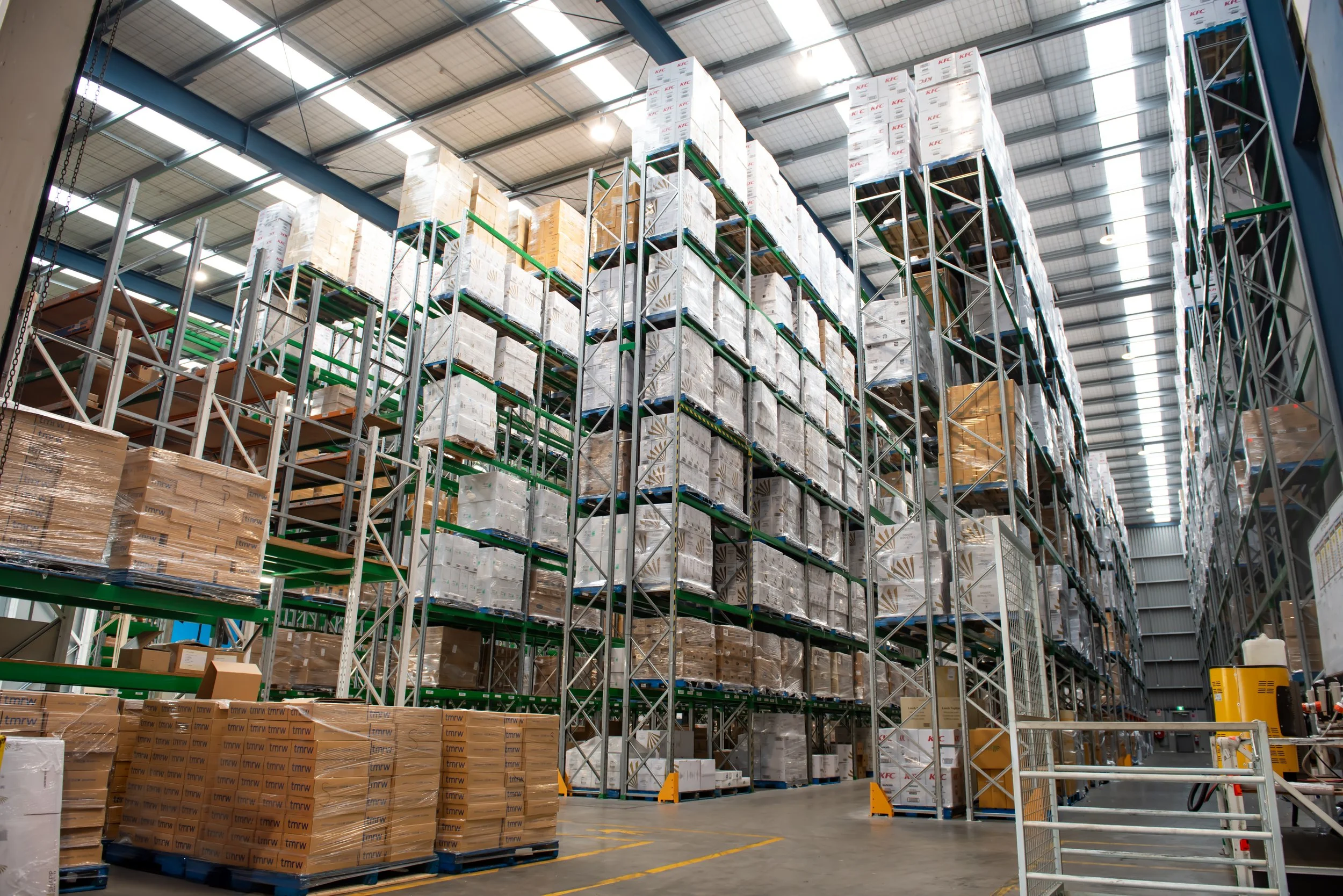 View of a warehouse with multiple shelves filled with boxes, pallets, and packages, including KFC branded boxes, under a high ceiling with metal beams.