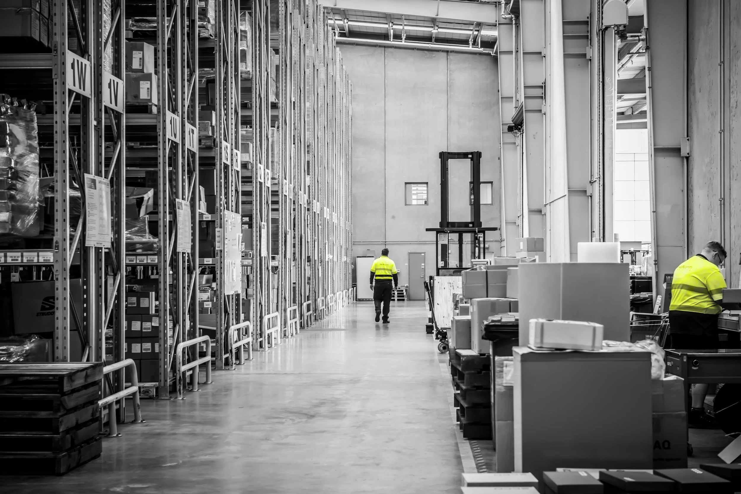 The image shows a large warehouse with shelves filled with boxes and packages. Two workers wearing yellow vests are inside, one walking through the aisle and another working at a desk. The warehouse has high ceilings and large windows allowing natural light.