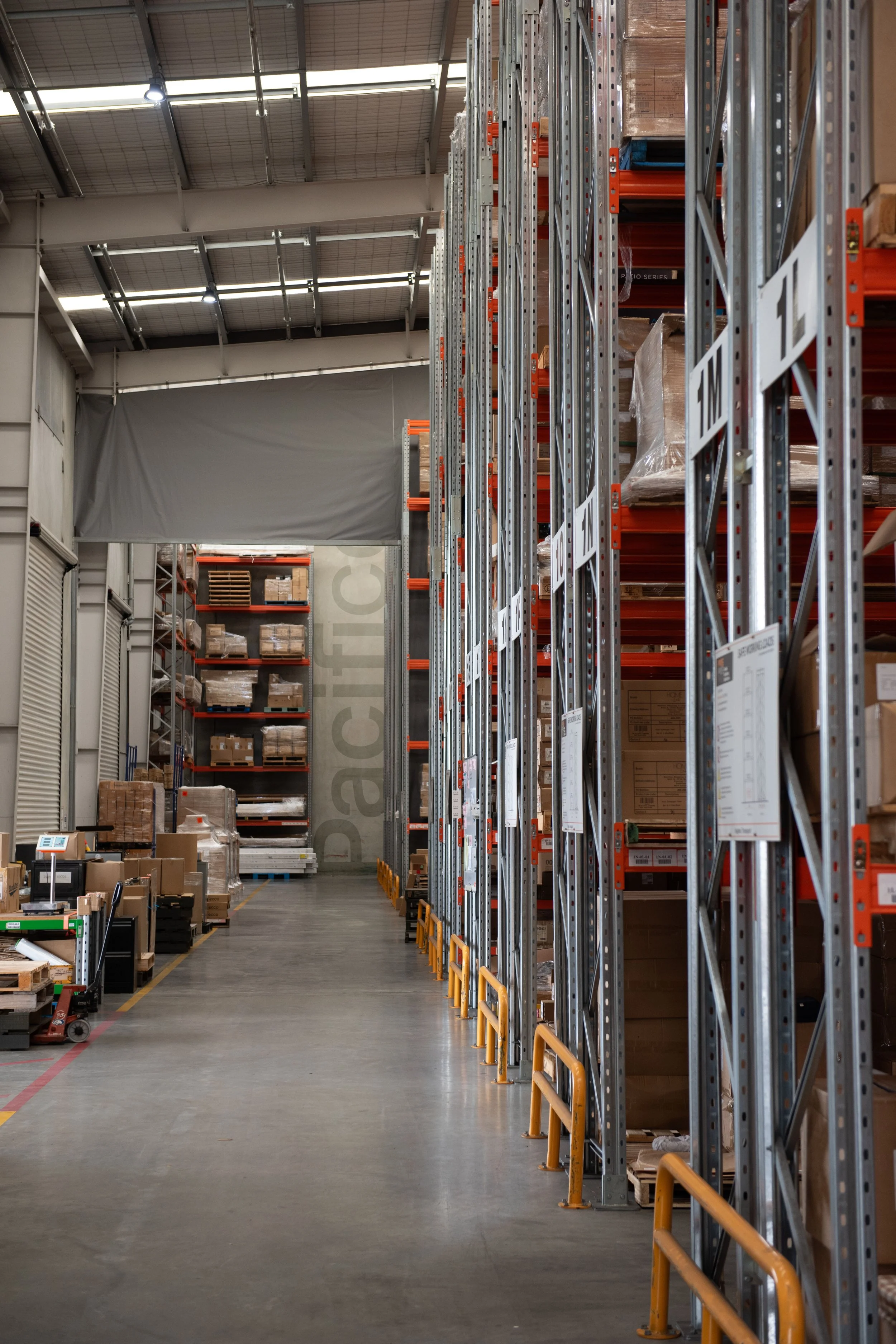 Warehouse aisle with tall metal shelving filled with boxes and packaging materials. The space has high ceilings, industrial lighting, and a smooth concrete floor.
