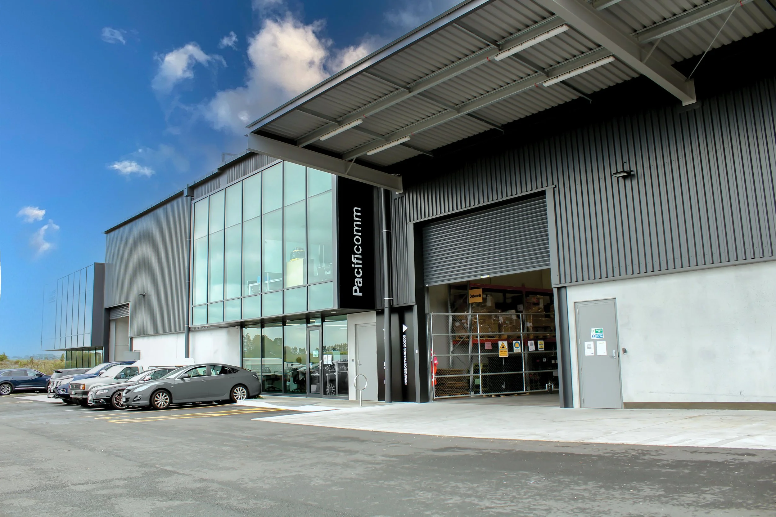 Exterior of a modern warehouse or distribution center with a large glass facade and a sign that reads 'Pacificflocamm.' There are parked cars in front, a large roll-up door, and a blue sky with clouds.