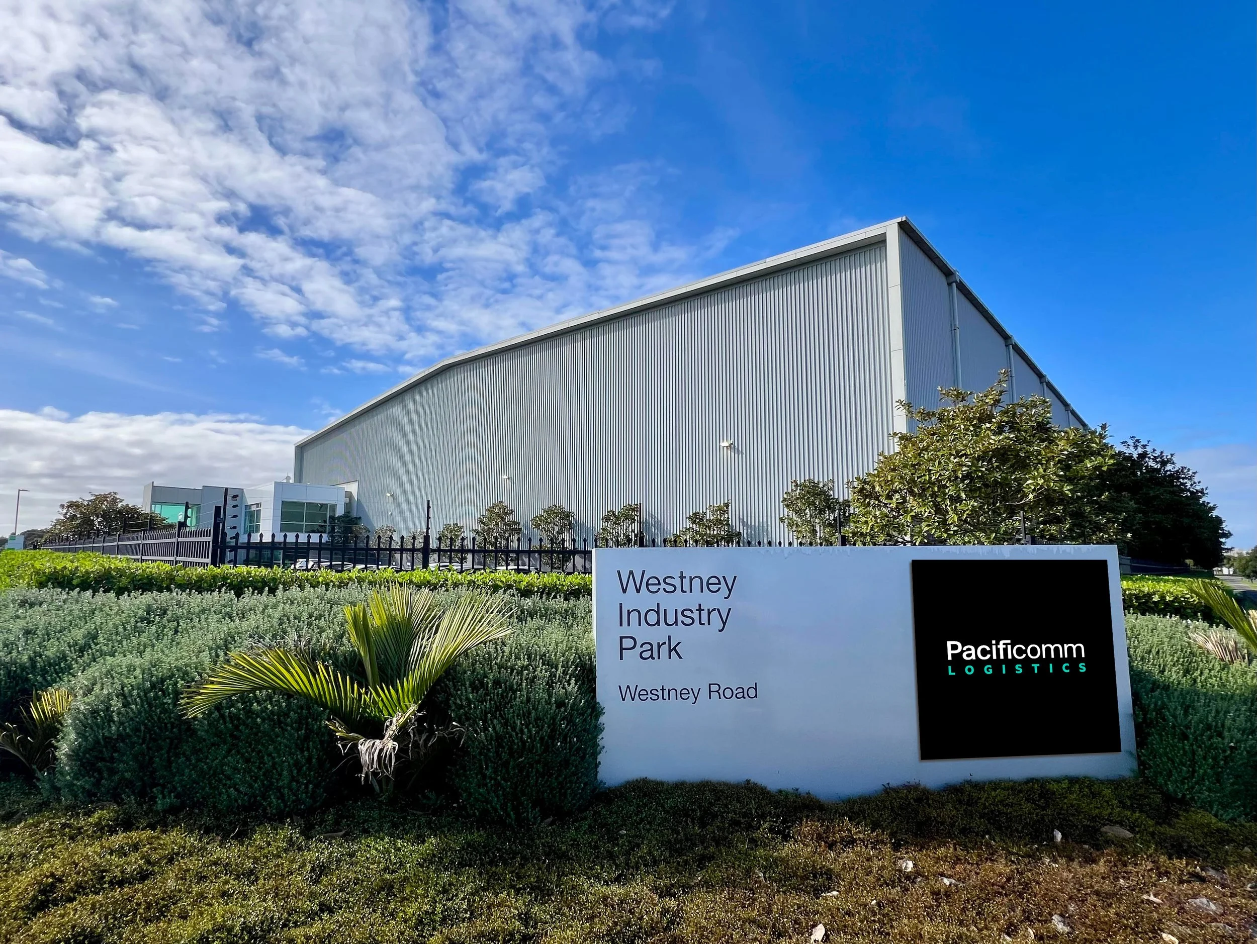 Sign for Westney Industry Park on Westney Road, with a large industrial building and landscaped bushes in front, under a partly cloudy sky.