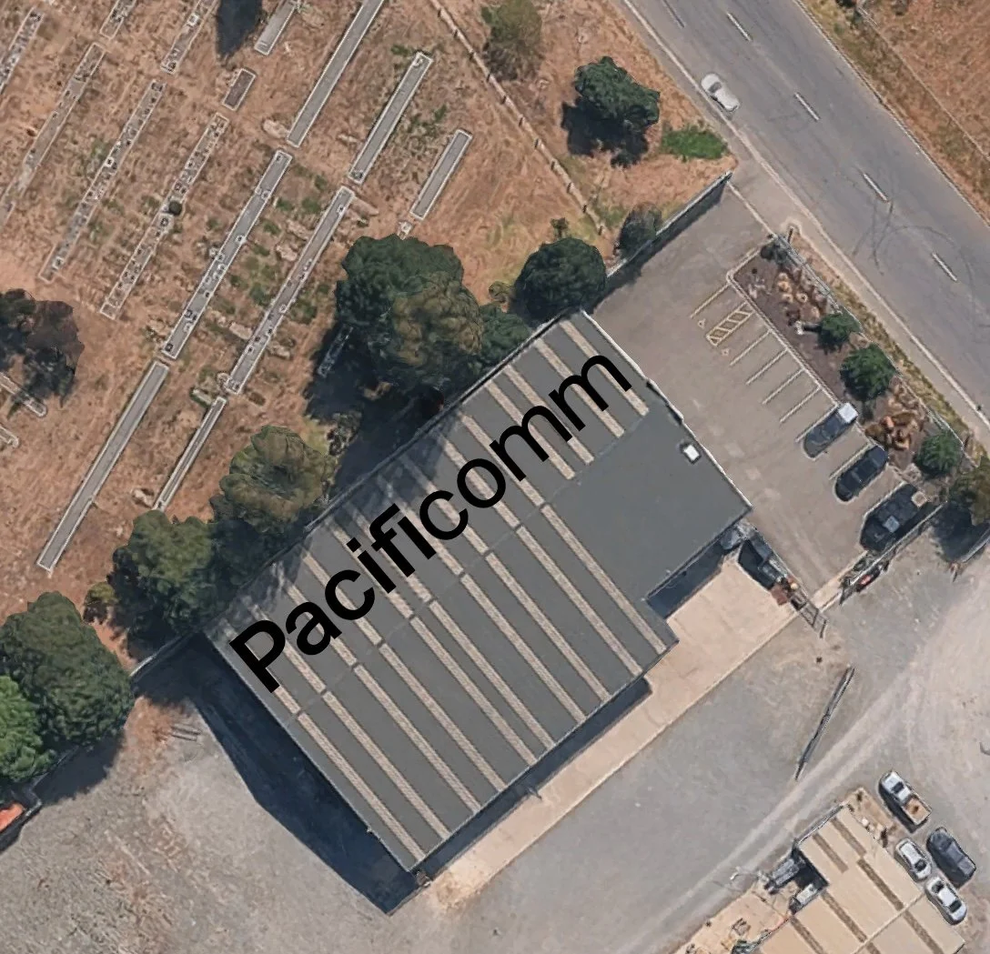 An aerial view of a building with a large roof and parking lot, adjacent to a road, with some trees and open space nearby. The roof has lines running across it, and the building and lot are part of a commercial or industrial area.