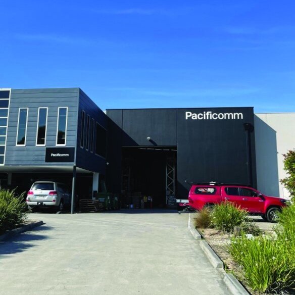 Exterior of a building with the sign 'Pacificcomm,' parked cars in front, and a clear blue sky.