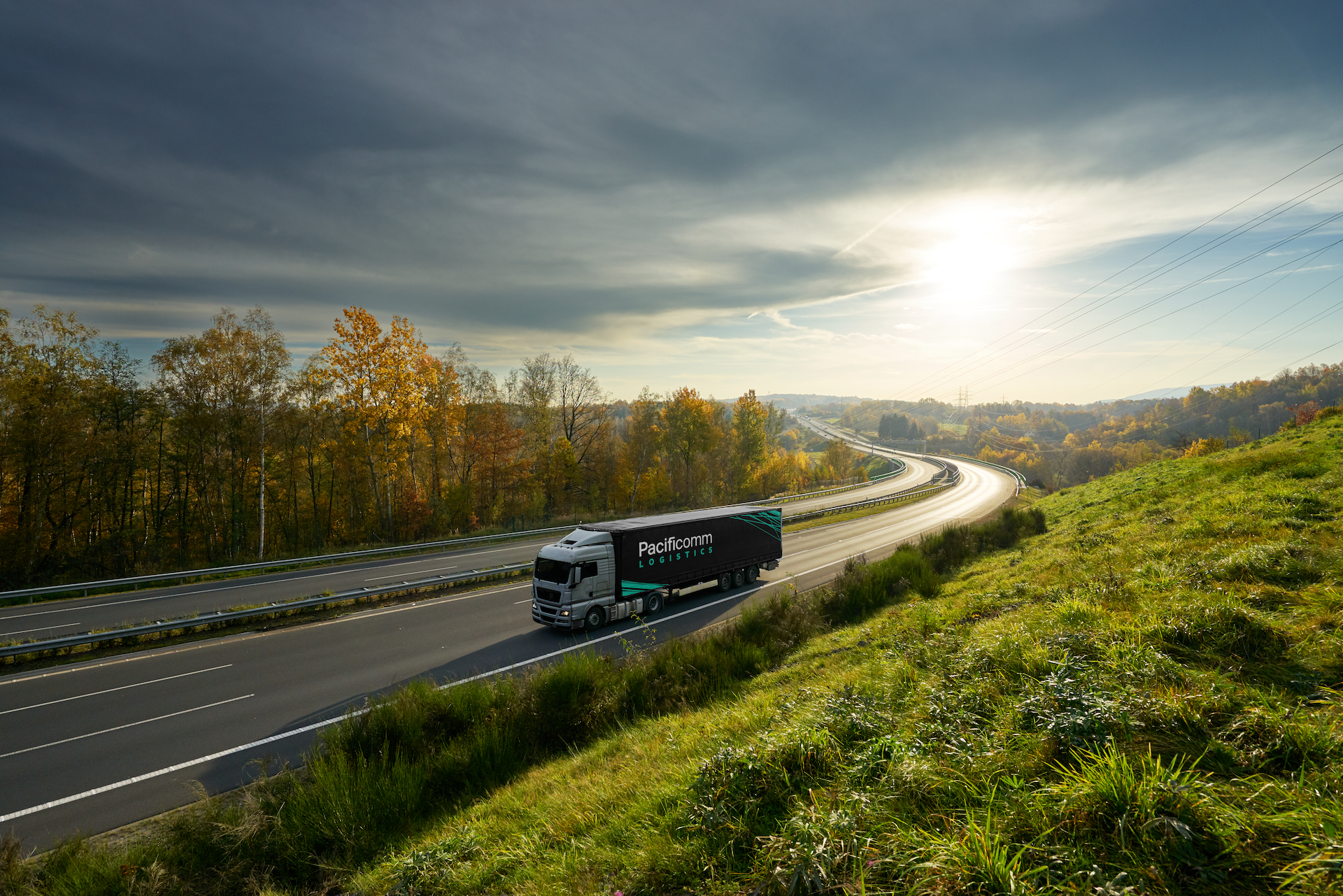 A Pacificcomm Logistics truck driving on a highway with green grass on the right and trees with fall foliage on the left, under a cloudy sky with the sun shining.