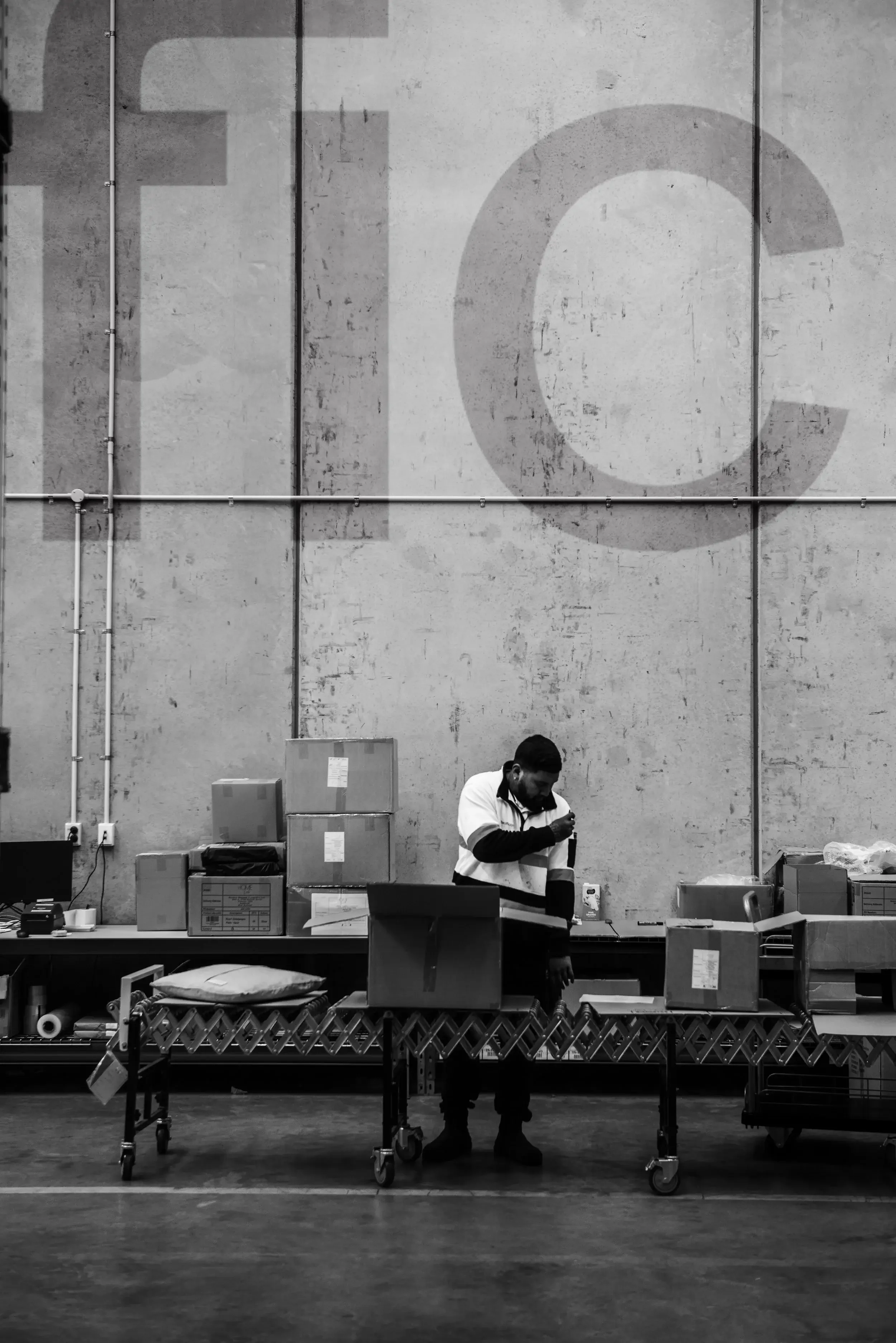 A warehouse or storage area with stacked boxes and a person working behind a table surrounded by more boxes. Large letters on the wall spell 'FC'.