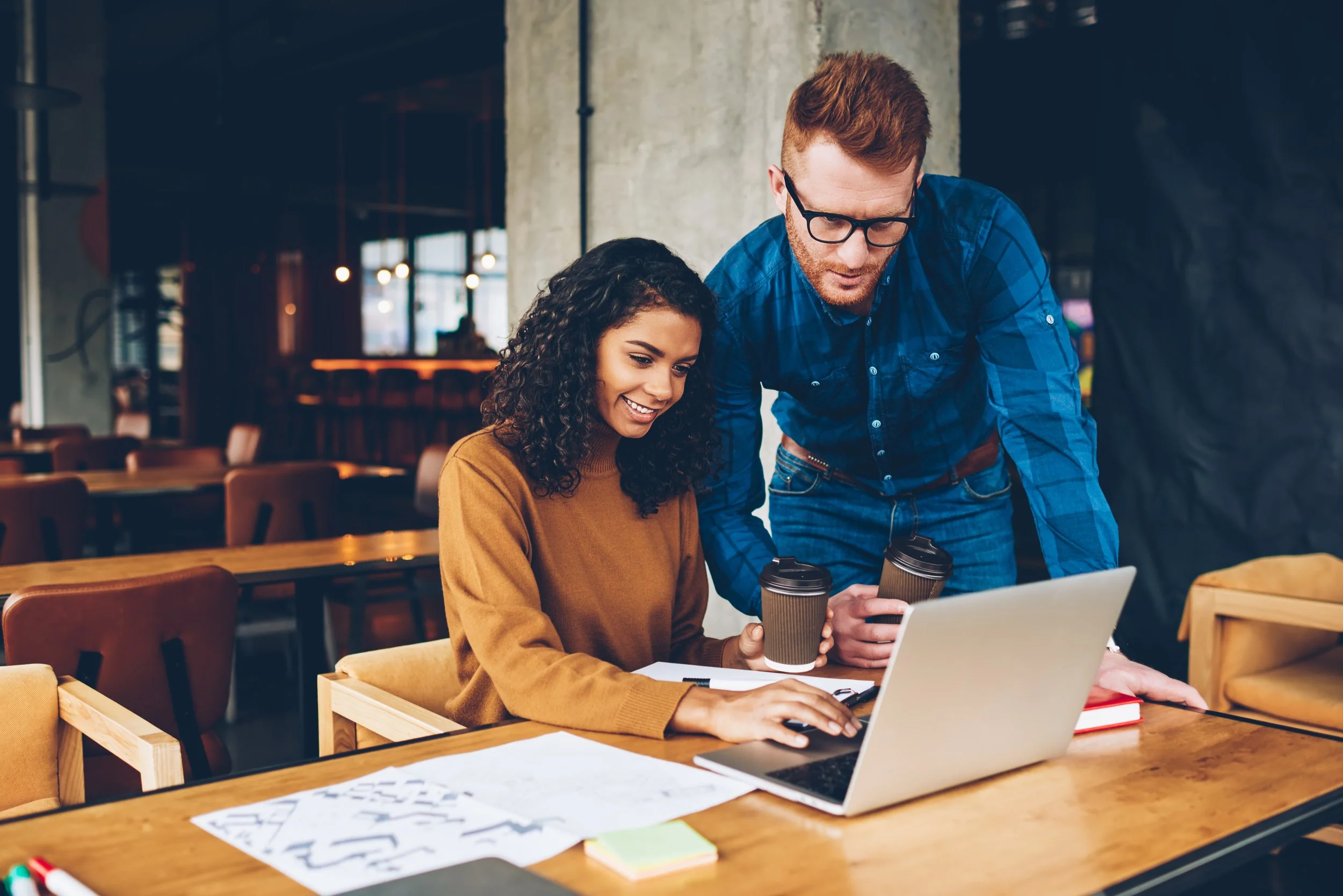 A woman with curly dark hair in a brown sweater working on a laptop at a wooden table in a cafe, holding a coffee cup, with a man with glasses and red hair standing beside her, also holding a coffee cup, both looking at the laptop screen.