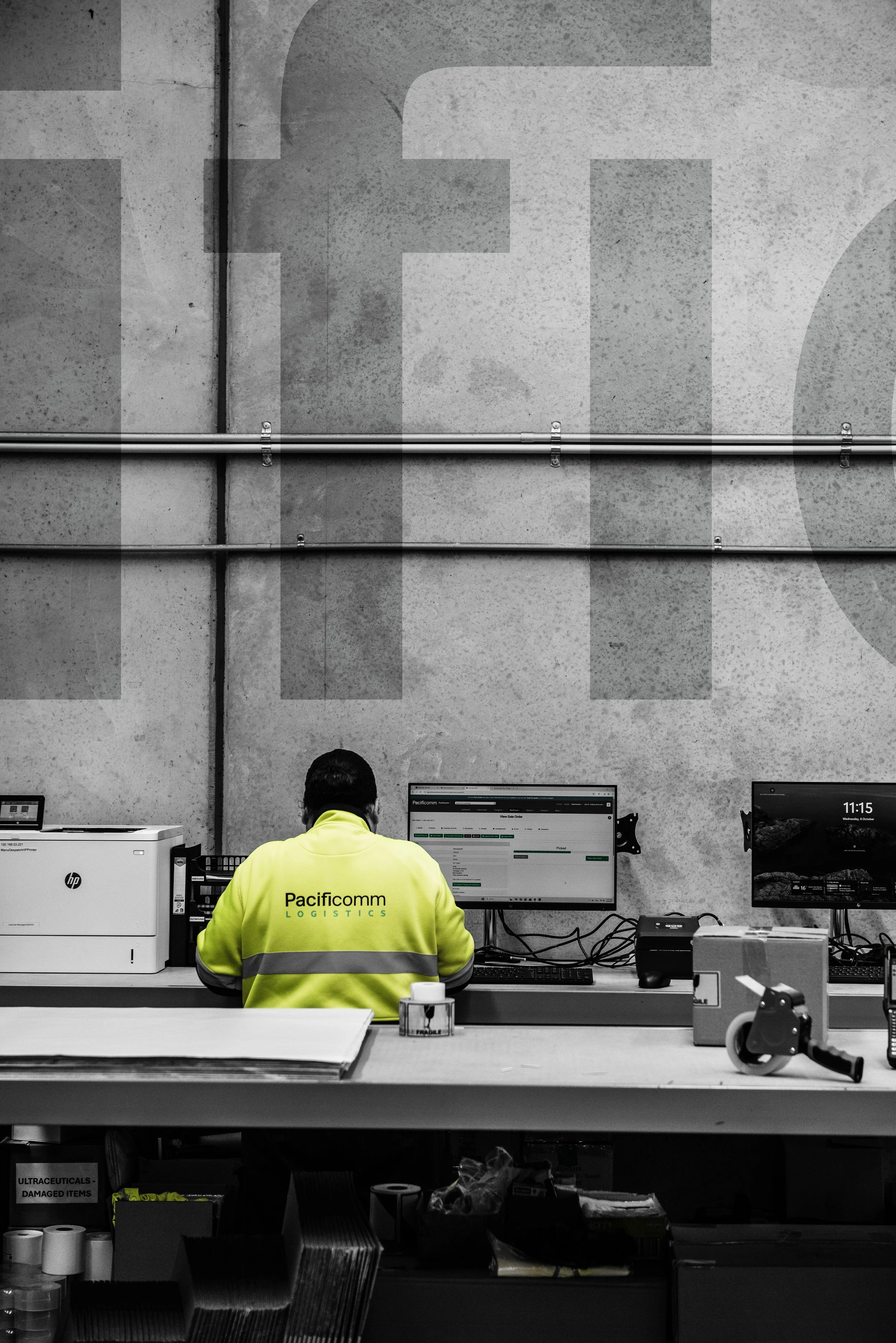 An employee wearing a yellow high-visibility jacket with 'PacifiComm Logistics' printed on the back, working at a desk with multiple computer monitors in an industrial or warehouse setting. The background features a concrete wall with large faded letters and exposed pipes.
