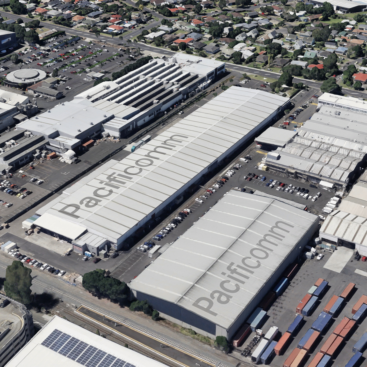 An aerial view of industrial warehouses and commercial buildings with parking lots and an adjacent residential neighborhood with single-family homes and greenery.