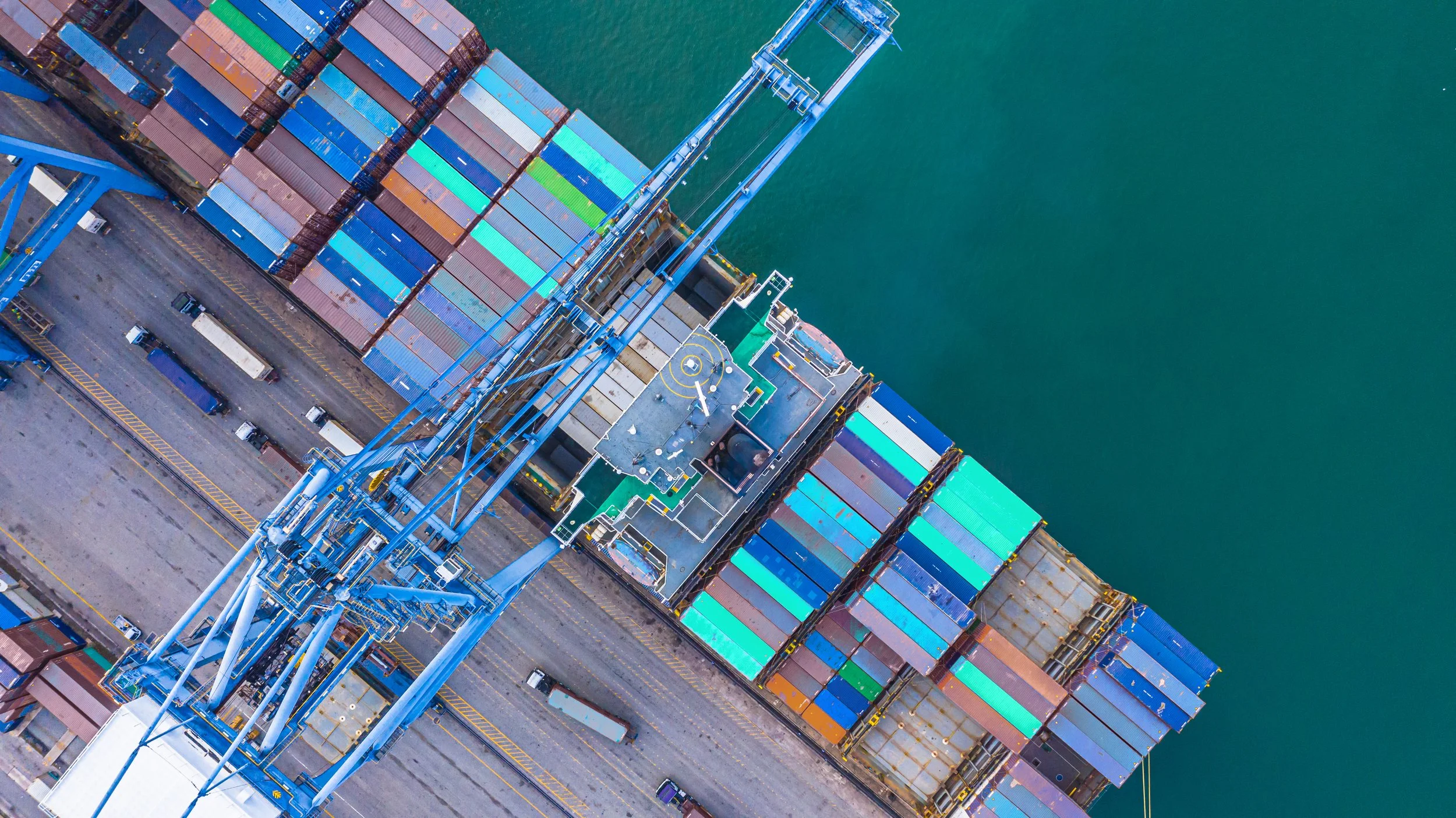 An aerial view of a shipping port with numerous stacked shipping containers in various colors, a large crane for loading and unloading, and water in the background.