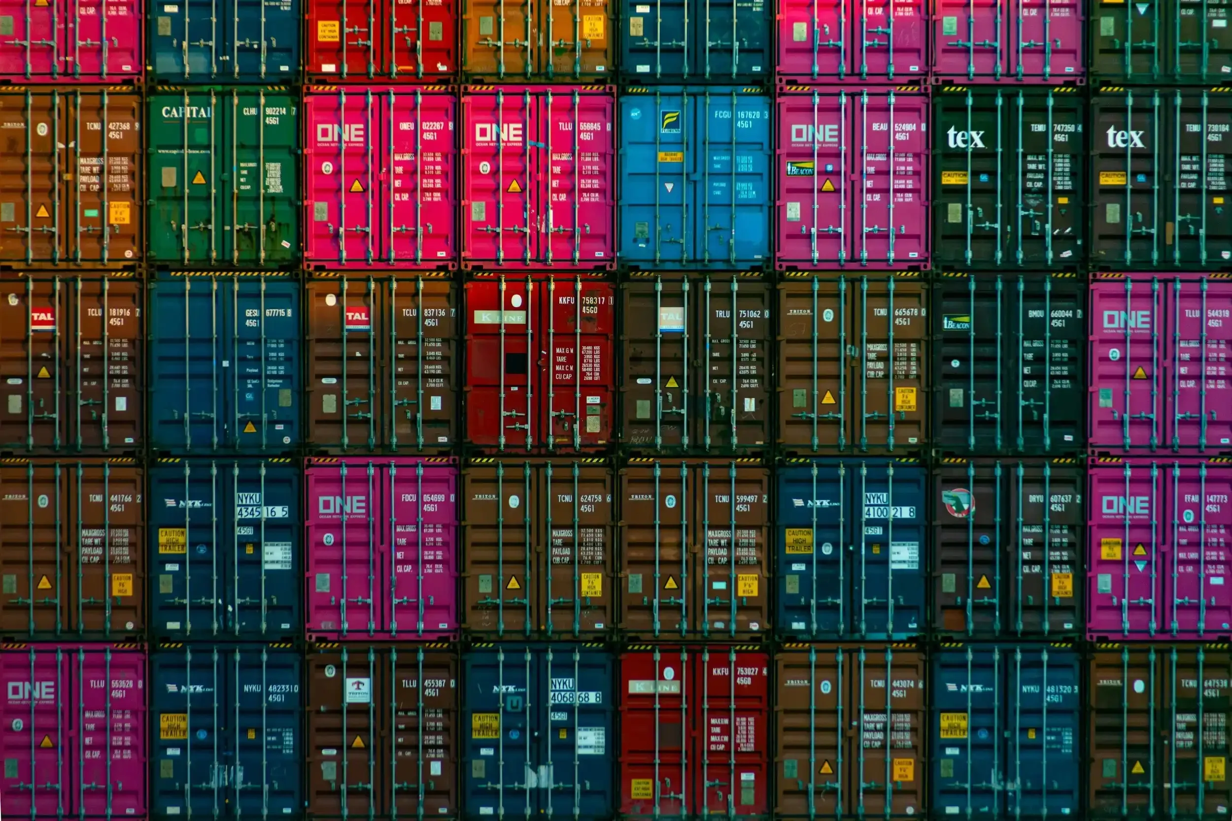 Stacked shipping containers in various colors including pink, blue, green, orange, and red, arranged in rows at a port.