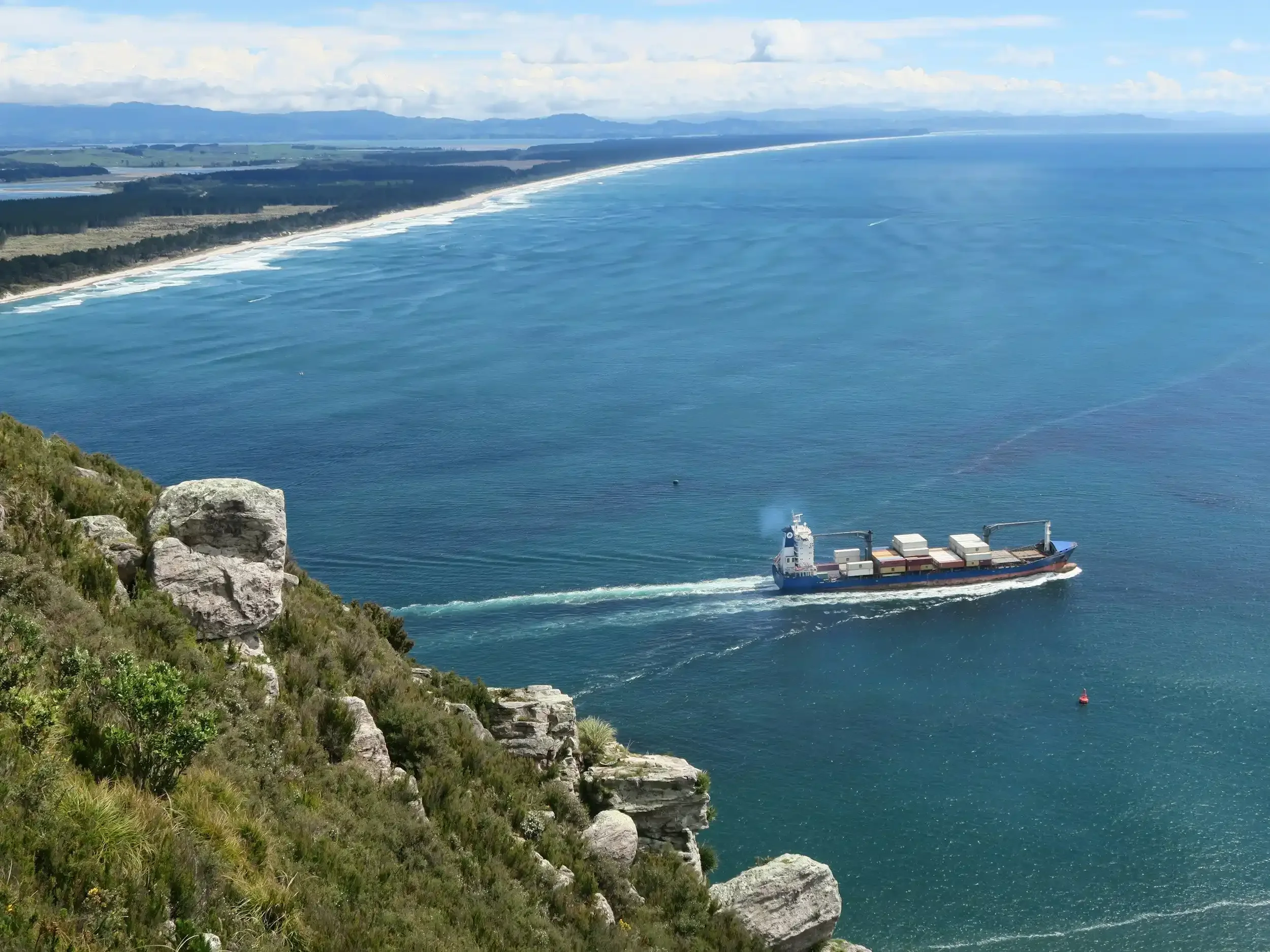 A large cargo ship sailing in the ocean near a rocky coastline with green bushes and trees, and distant land in the background.