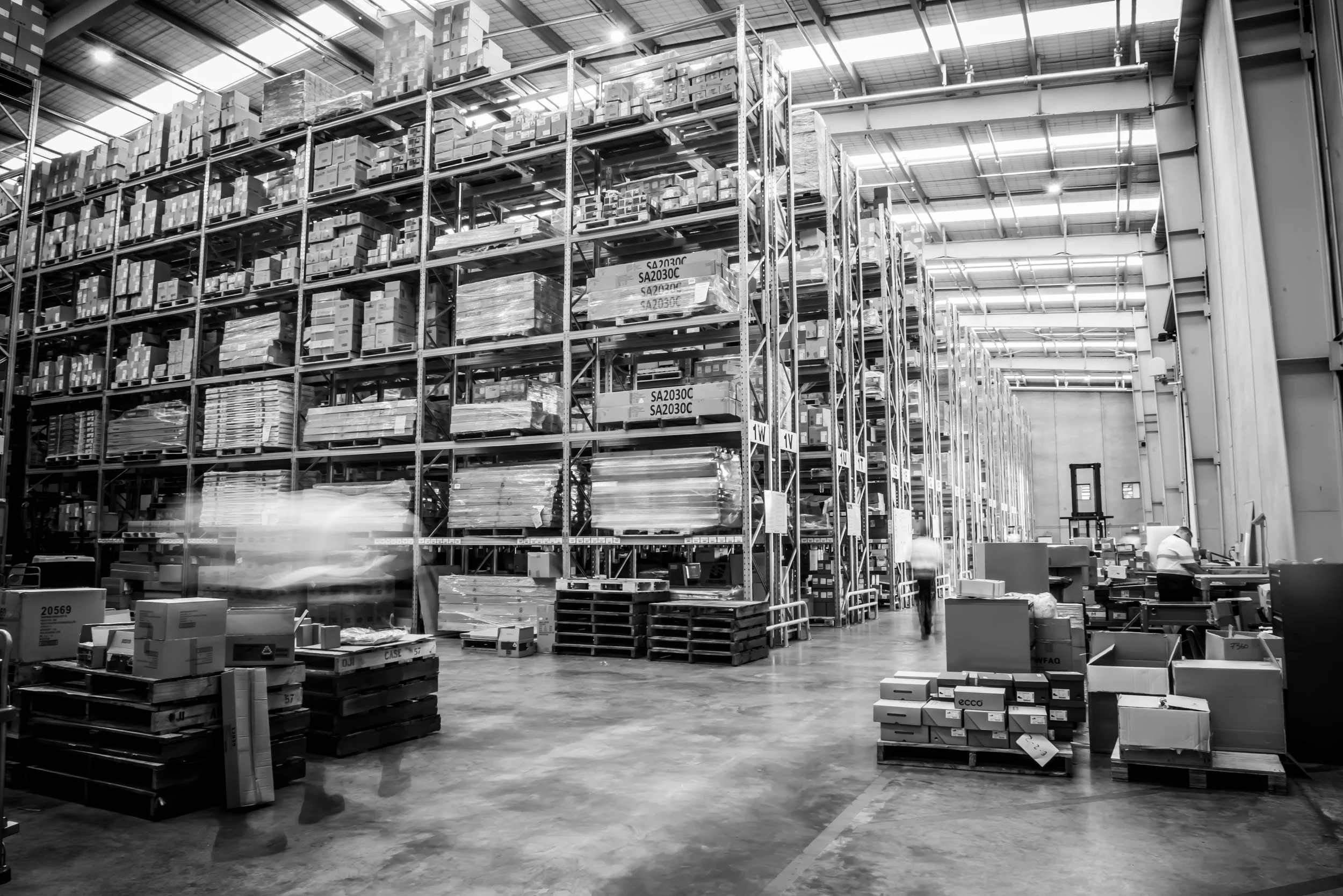 Interior of a large warehouse with tall shelves stocked with boxes and pallets, and a worker moving items in the foreground.
