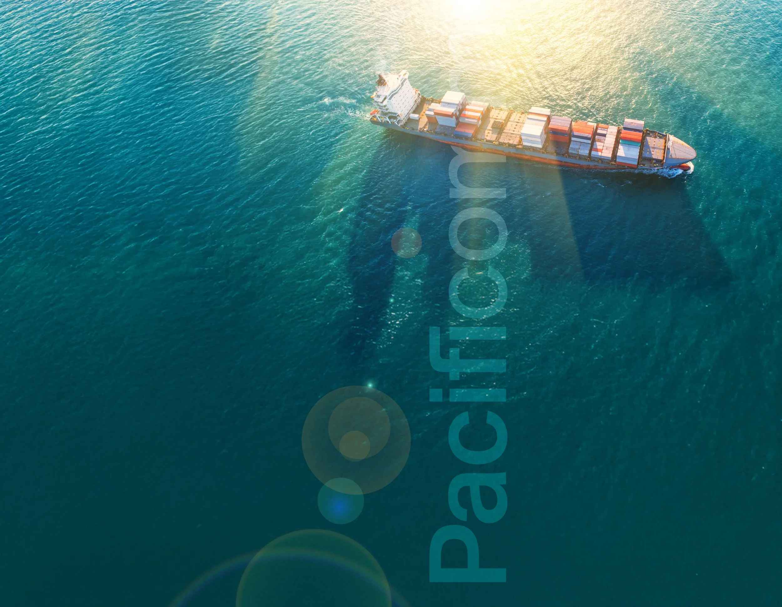 An aerial view of a cargo ship with stacked containers sailing in the ocean near the coast, illuminated by sunlight.