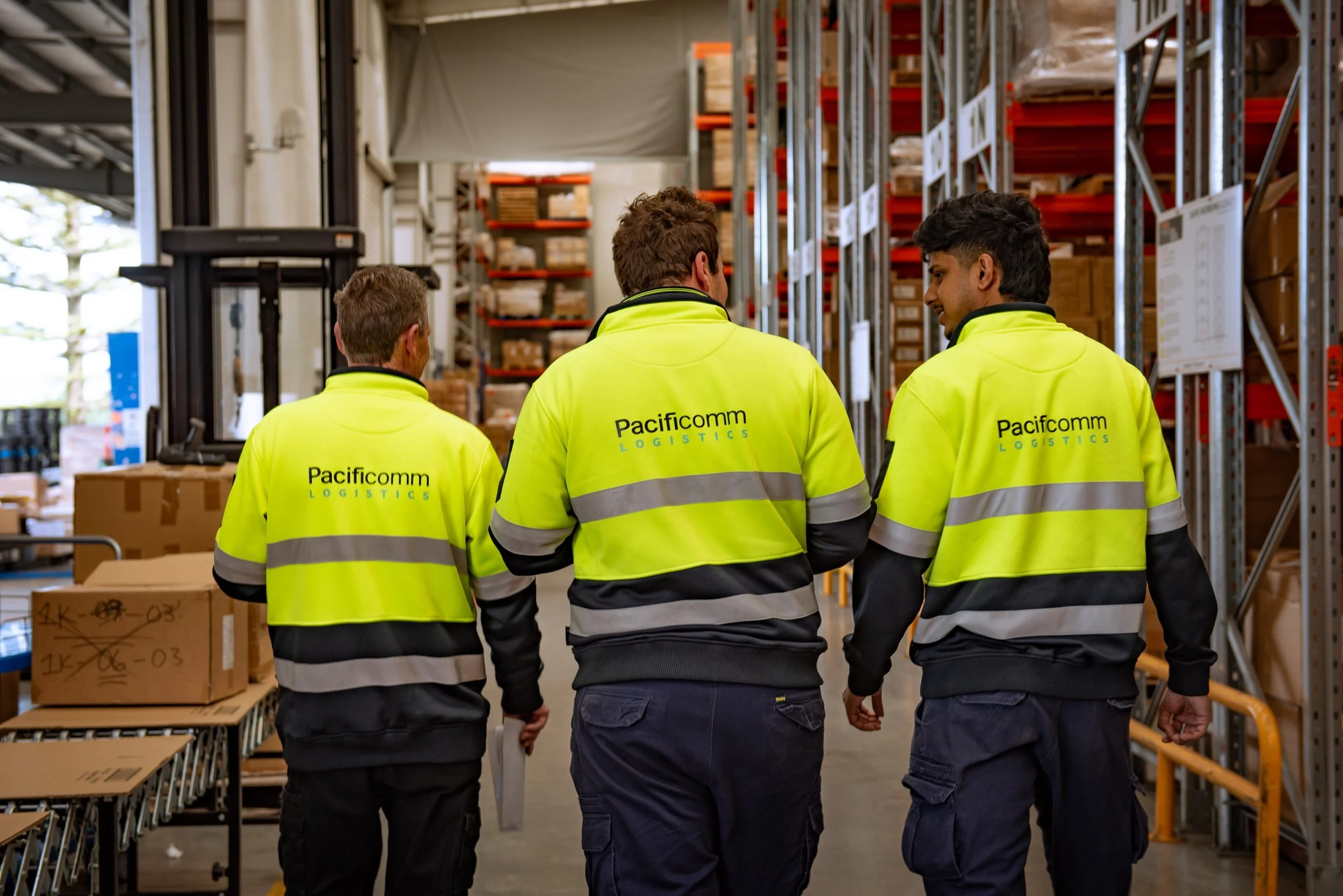 Four warehouse workers wearing bright yellow high-visibility jackets with 'Pacificcomm LOGISTICS' printed on the back, standing together and talking inside a storage facility with shelves filled with boxes.