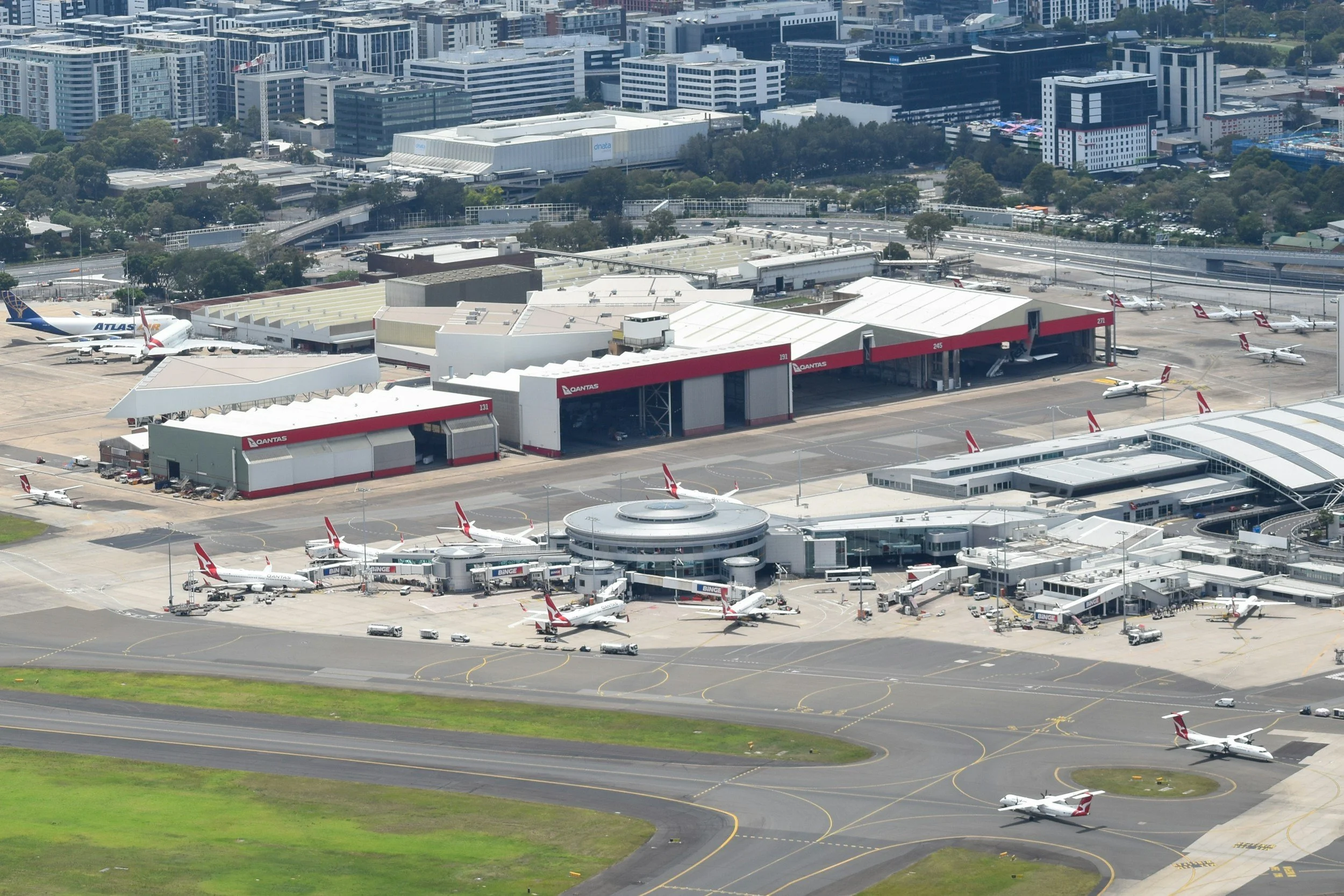 Aerial view of an airport with airplanes parked at gates, terminal buildings, and surrounding cityscape with tall buildings and green areas.