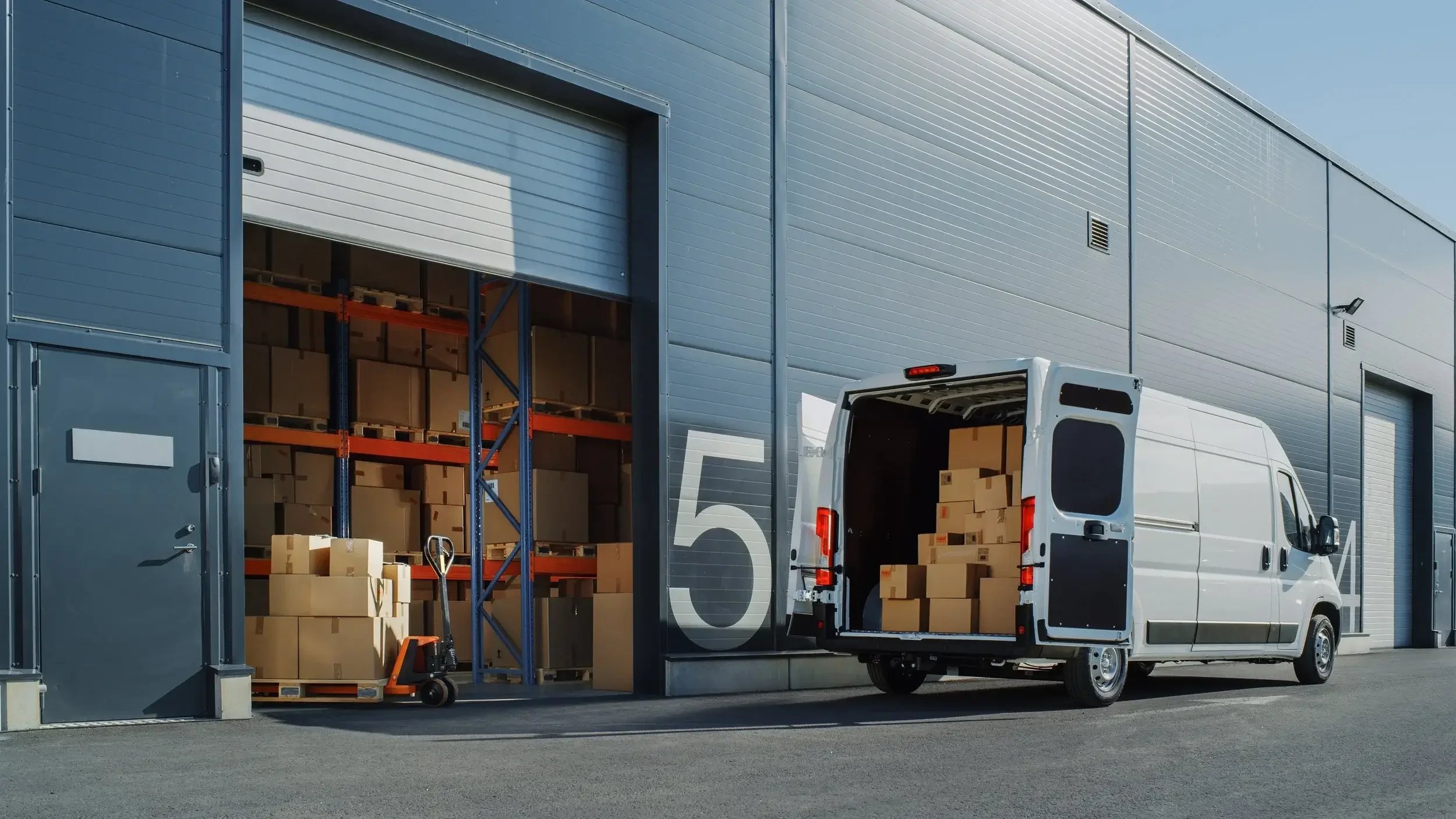 A white delivery van backed into a warehouse with brown boxes inside and outside. The warehouse has large roll-up doors and a blue exterior.