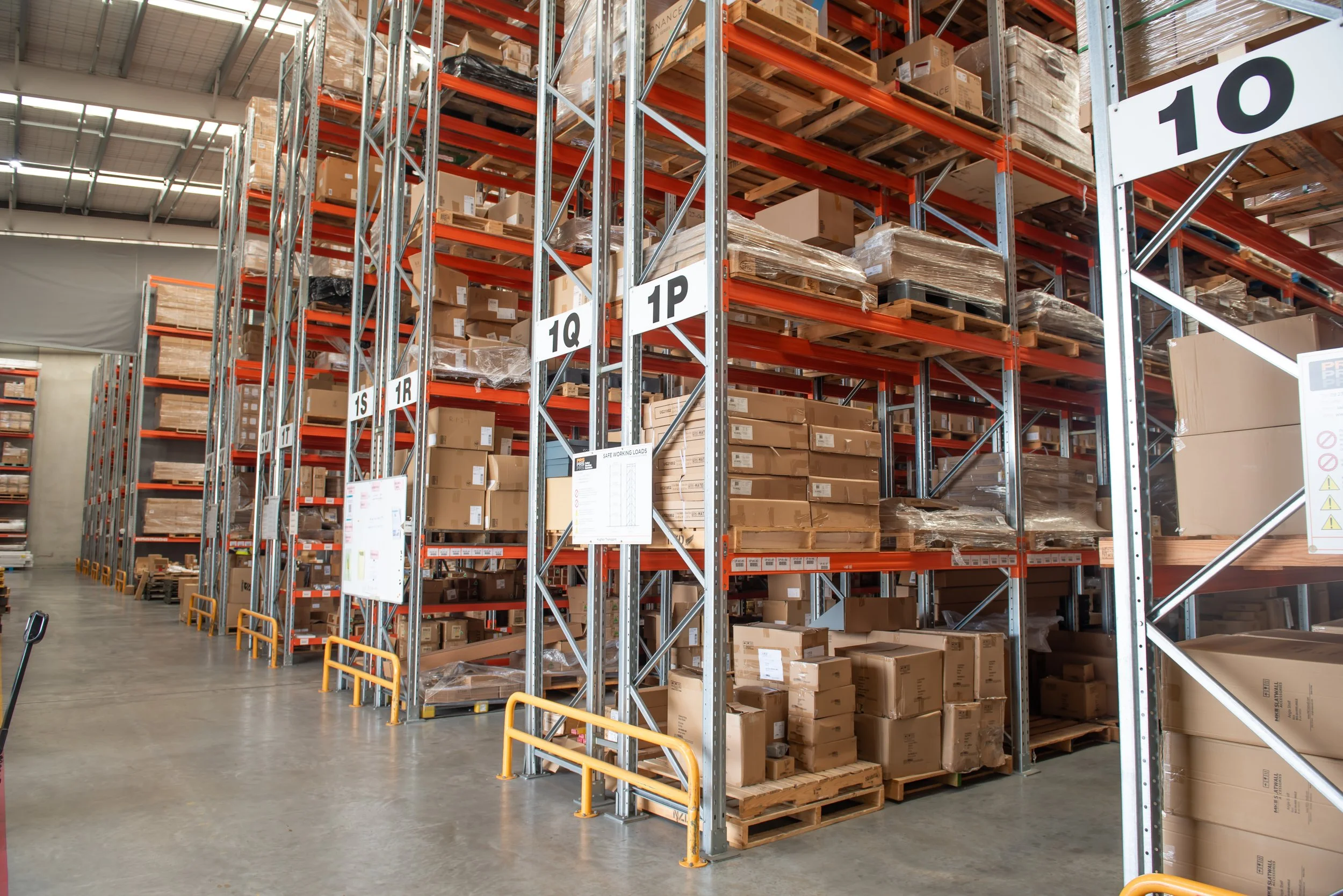 Warehouse aisle with tall orange and gray metal shelves filled with numerous cardboard boxes, some wrapped, arranged in various levels. Signage indicates aisle numbers.