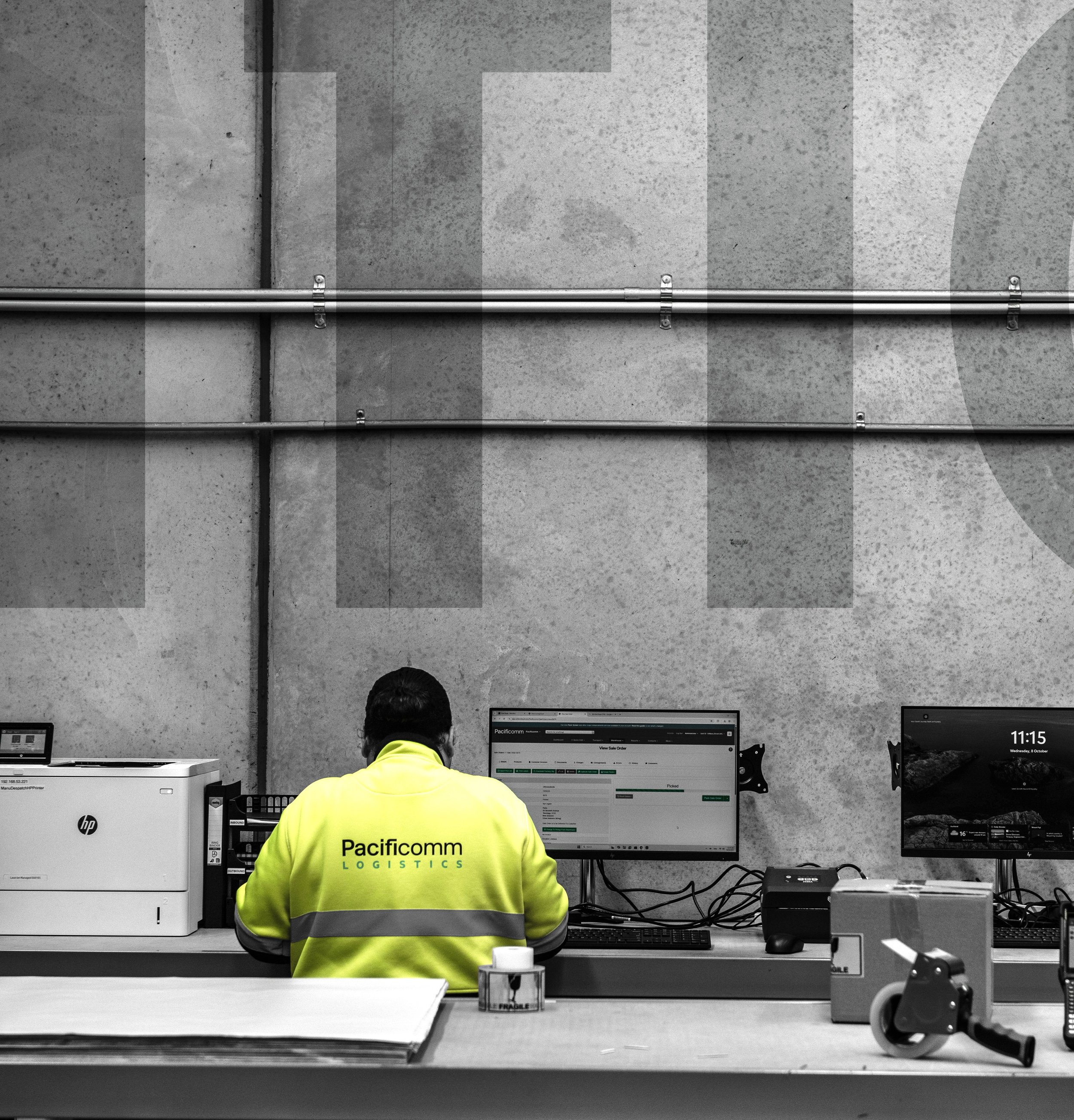 A person in a yellow jacket with the words "Pacificomm Logistics" on the back is working at a desk with multiple computer monitors. The room has a concrete wall with metal piping and various office supplies on the desk.