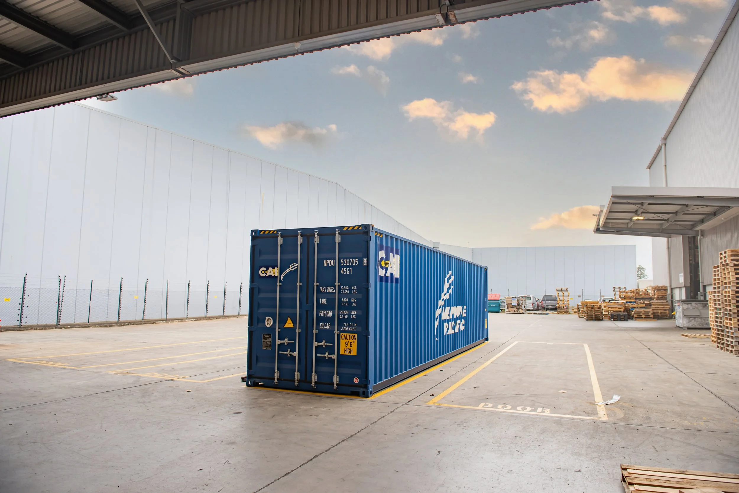 Empty parking lot with a large blue shipping container and warehouse buildings, some pallets and parked cars under a partly cloudy sky.