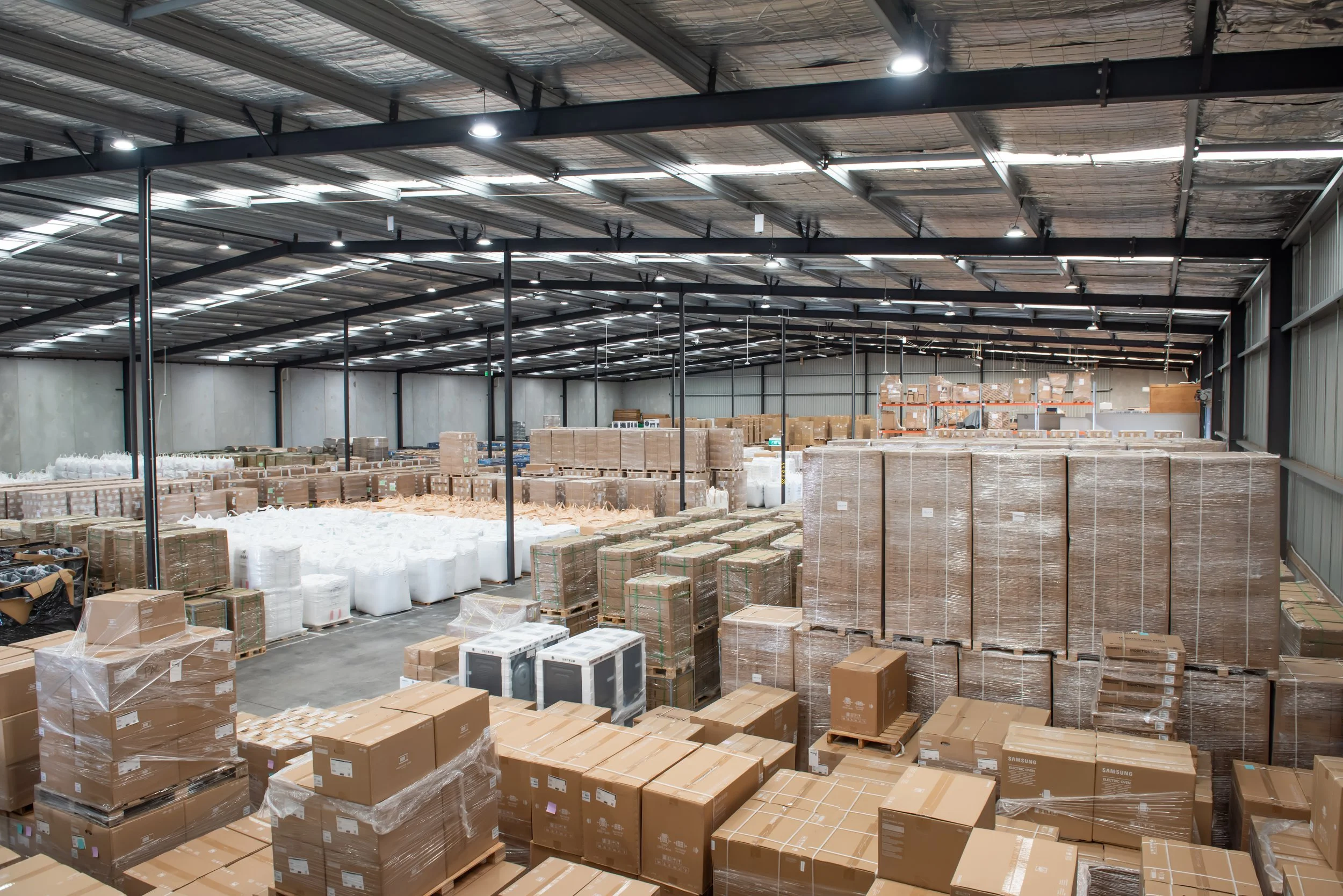 Inside a large warehouse with metal walls and a high ceiling, stacked with numerous cardboard boxes on pallets, some wrapped in plastic, and various other warehouse items.