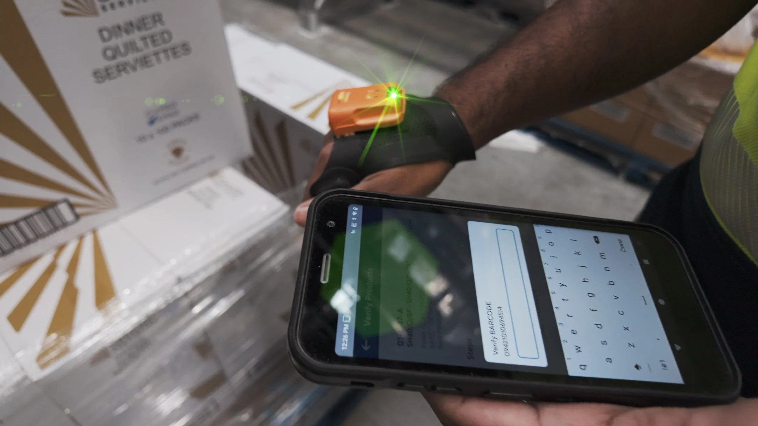 Person scanning a barcode with a handheld scanner wearing a wristwatch, while a mobile phone with a keyboard is held in their other hand.