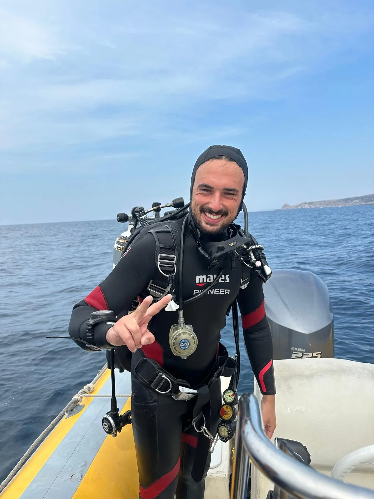 Uomo sorridente con attrezzatura subacquea su barca vicino al mare, facendo segno di pace