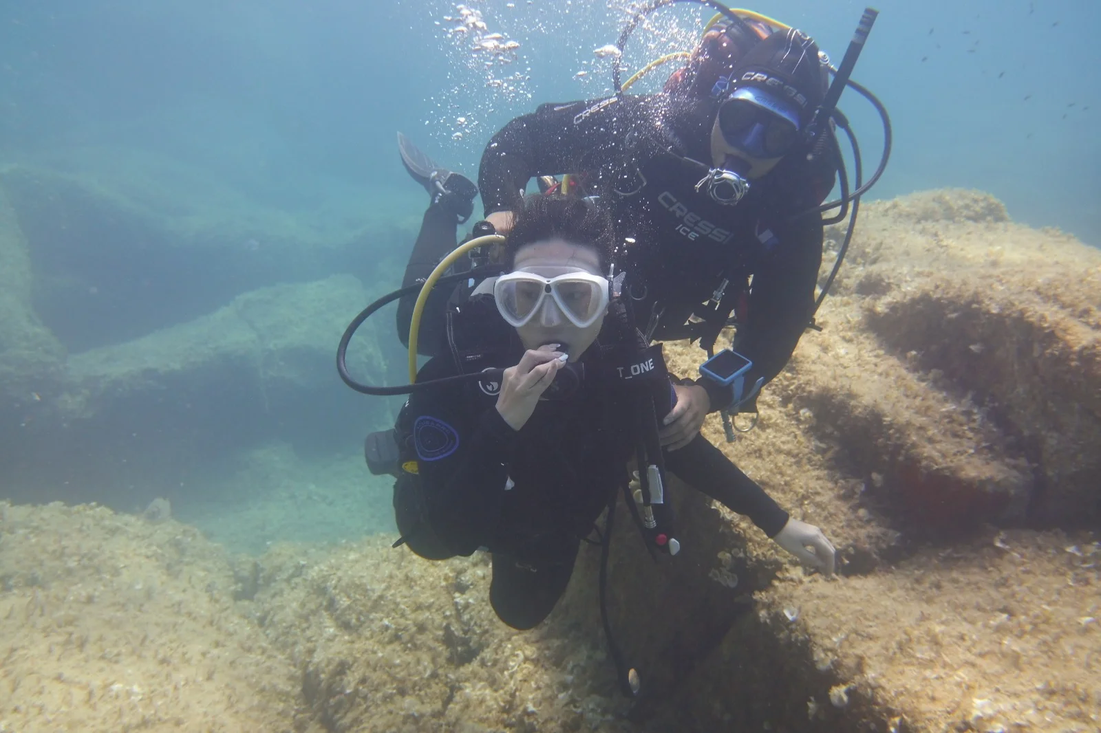 Due subacquei in immersione tra scogli e fondo marino sabbioso, uno dei quali in primo piano che cattura qualcosa con la mano, entrambi con maschere e attrezzature da immersione.