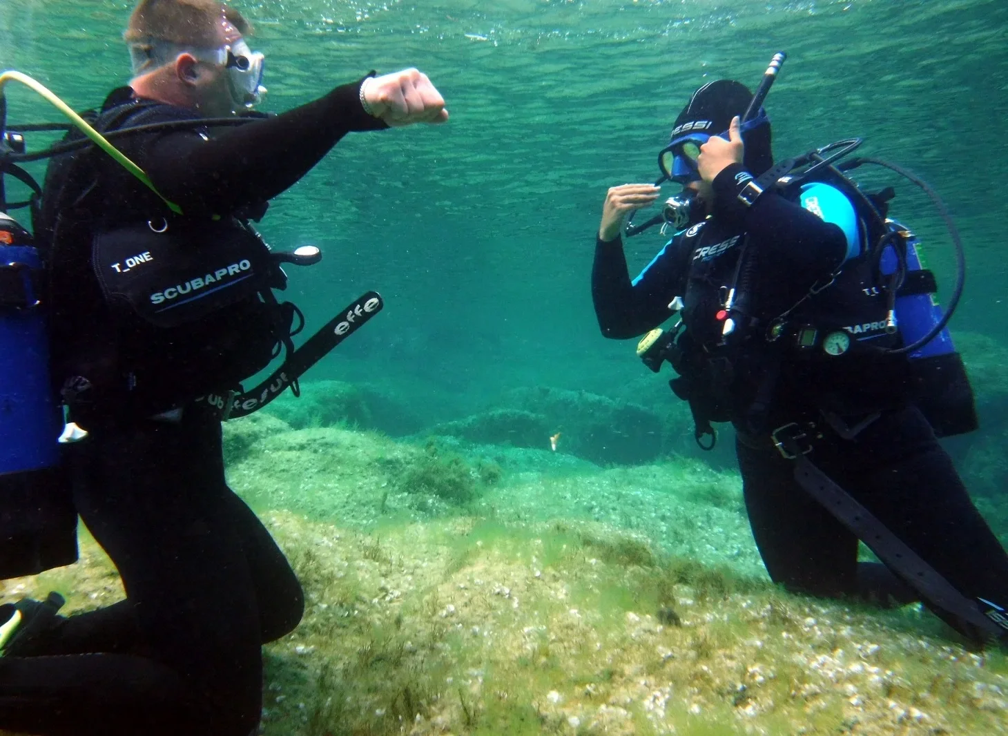 Due subacquei in immersione, uno con il pugno chiuso e l'altro che saluta, in acqua vicino al fondo sabbioso con alghe marine.