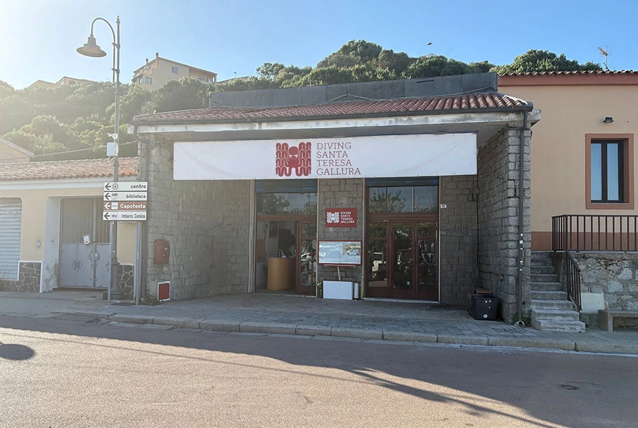 Edificio con segnaletica per diving Santa Teresa Gallura, in una zona urbana con colline sullo sfondo, giorno soleggiato.