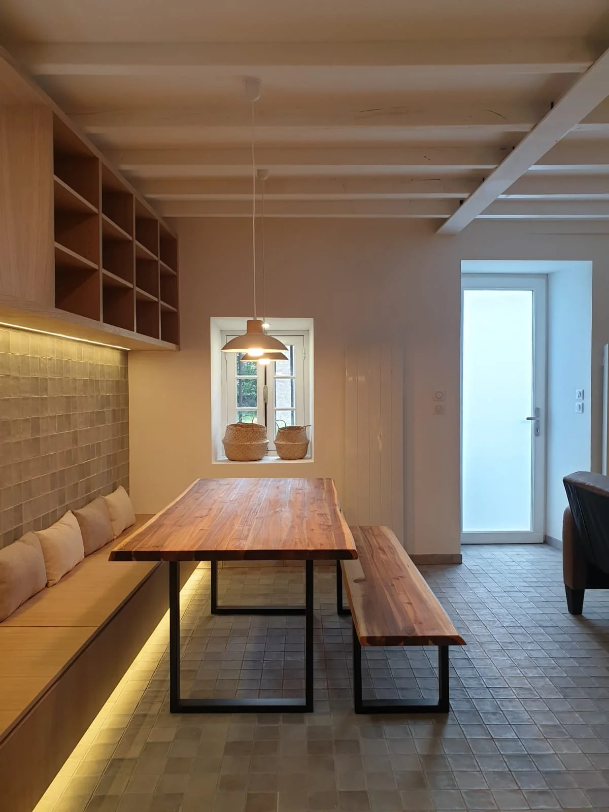 Cuisine moderne avec table en bois massif, niches en bois au mur, mur en carrelage beige, fenêtre avec paniers en osier et porte en verre opaque.